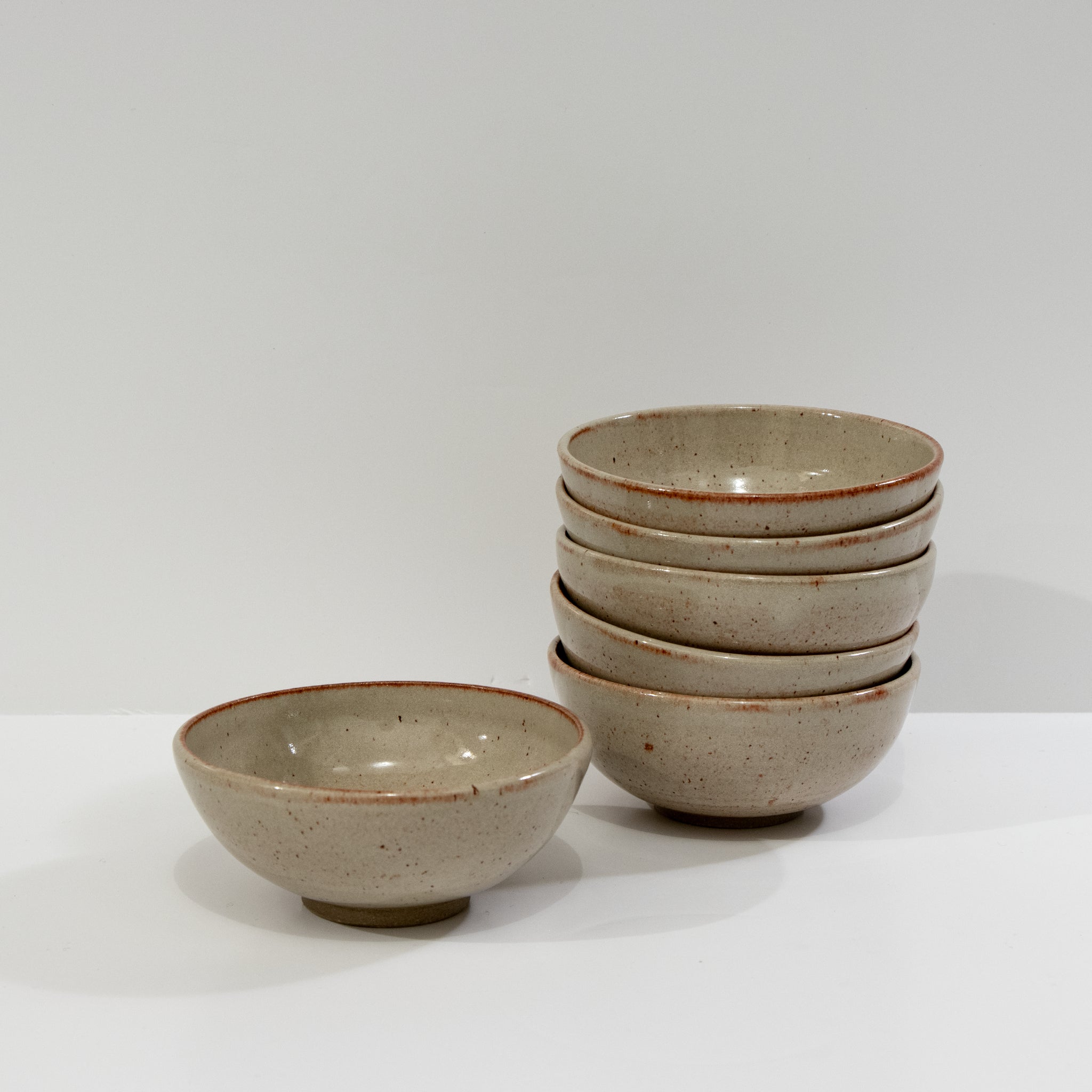 Zes AB Ceramics Mini Kom Geel Beige D10,5 kommen met een gespikkeld beige glazuur en bruine randen worden getoond; vijf staan er rechts opgestapeld en één staat apart links tegen een effen witte achtergrond.