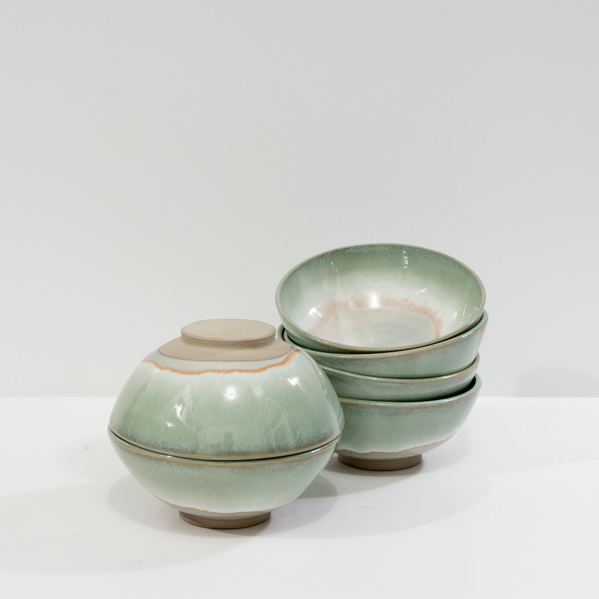 Vier AB Ceramics Mini Kom Wit Opaal/Lichtgroen D10,5 schaaltjes met een groen glazuur en bruine rand zijn afgebeeld op een witte achtergrond. Drie zijn gestapeld; één staat er ondersteboven naast, wat de unieke stijl van AB Ceramics benadrukt.