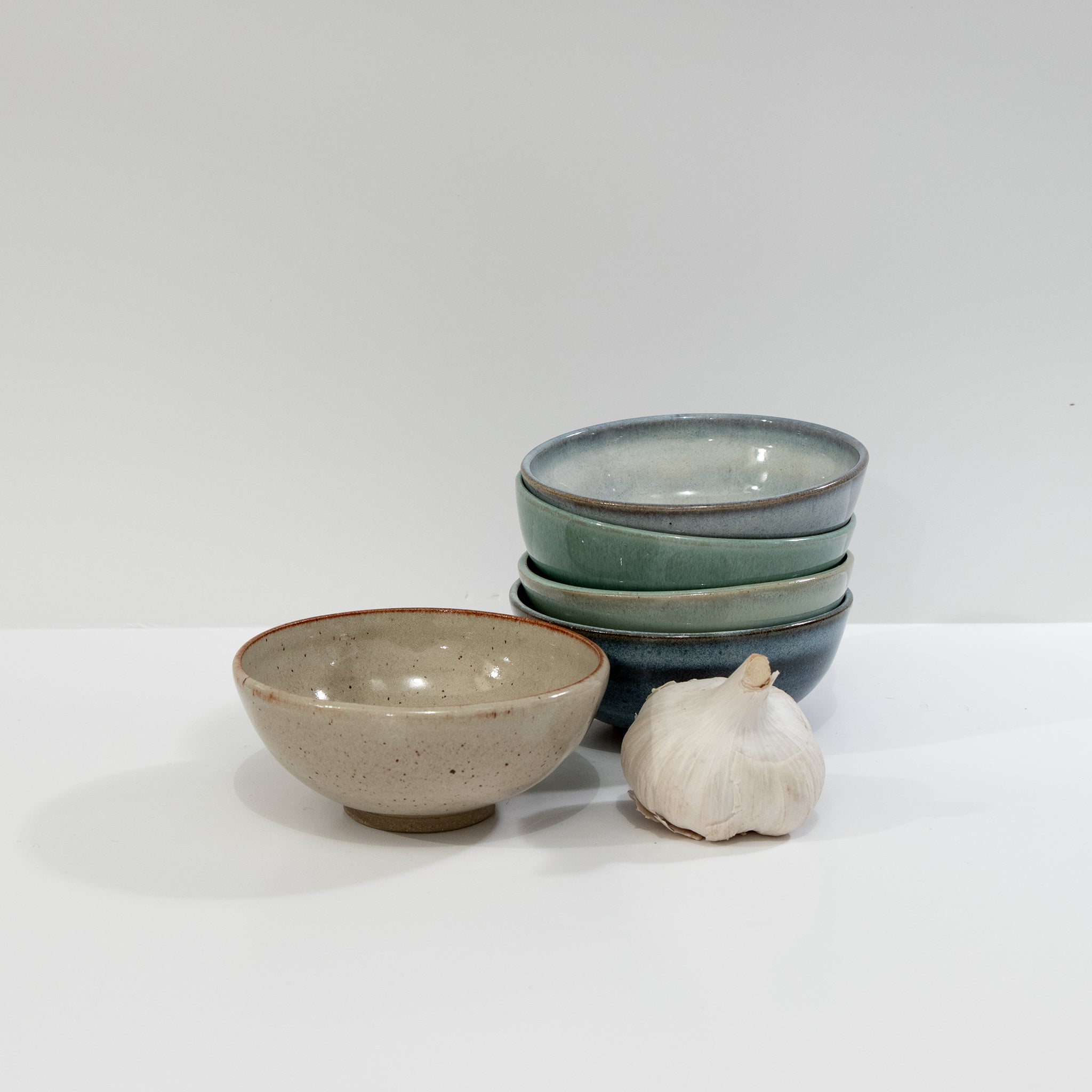 Drie AB Ceramics Mini Kom Rood Opaal D10,5 kommen zijn gestapeld, met een andere ernaast op een wit oppervlak; een hele knoflookbol is ook aanwezig.