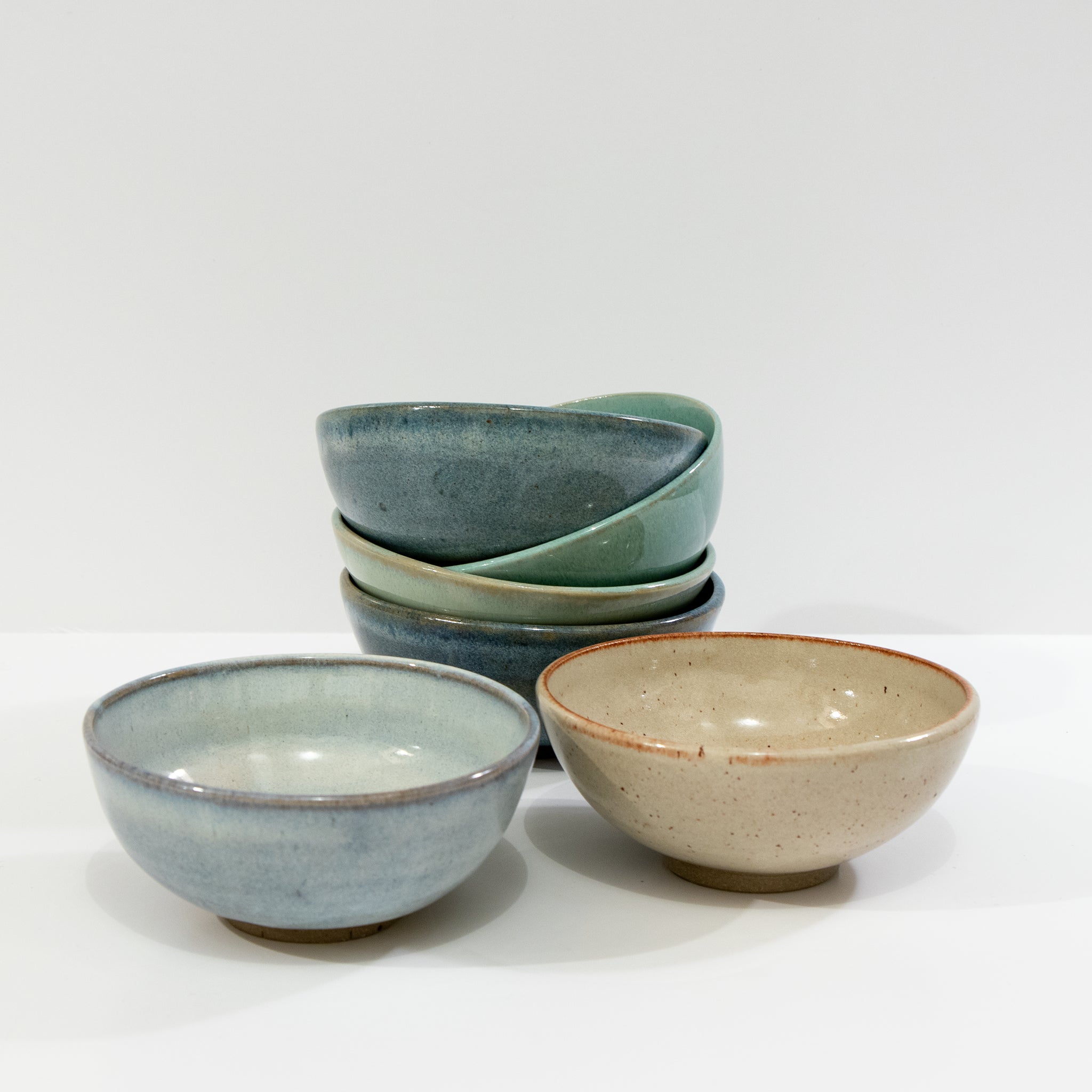 Vier AB Ceramics Mini Kom Geel Beige D10,5 keramische kommen zijn gerangschikt op een wit oppervlak - twee gestapelde blauwgroene kommen aan de achterkant, een lichtblauwe en een beige aan de voorkant - waarmee AB Ceramics' handgemaakte servies van steengoed klei wordt benadrukt.