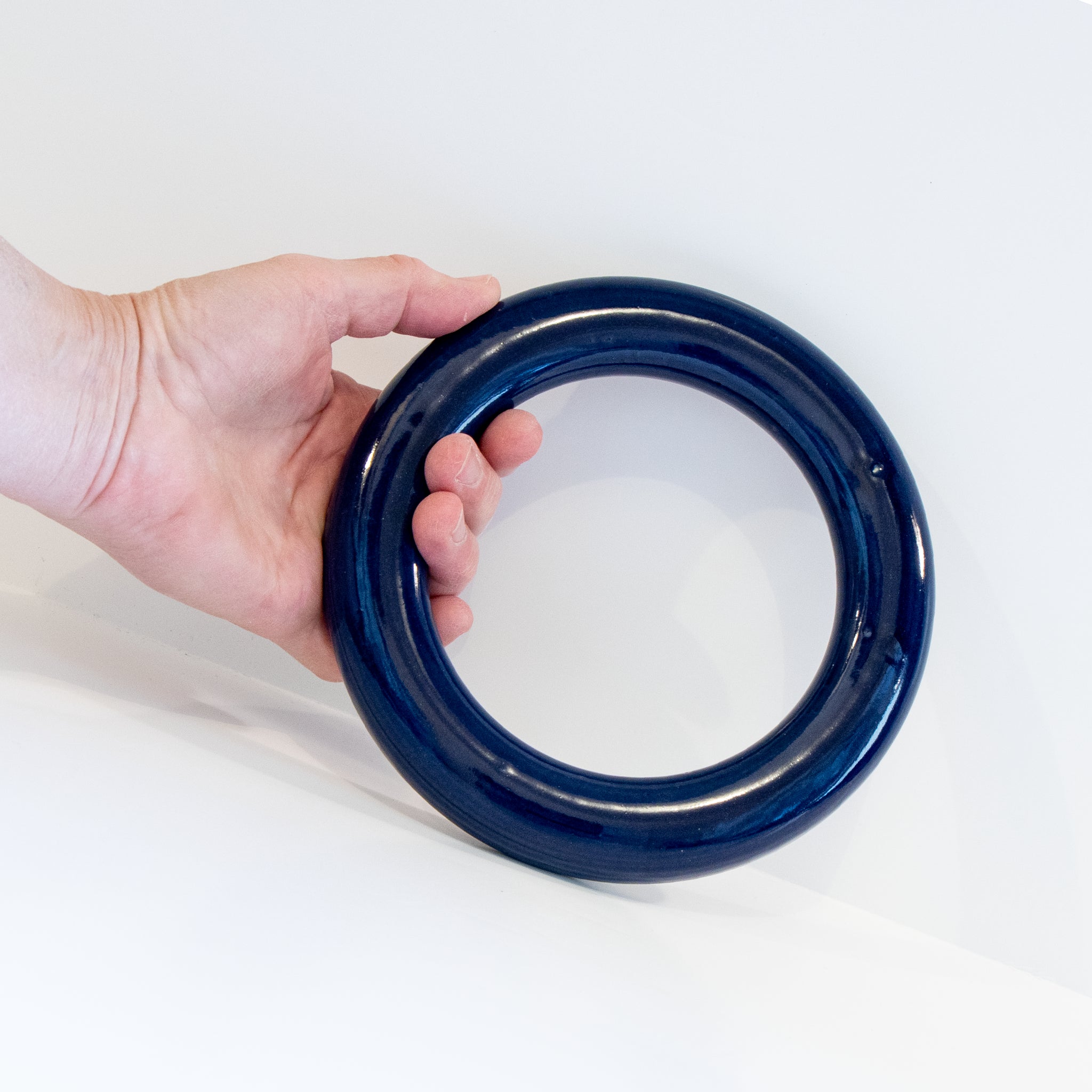 Een hand houdt de AB Ceramics Pot Onderzetter Blauw D16,5 vast, met een glanzend donkerblauw glazuur en een minimalistisch ontwerp van AB Ceramics, tegen een witte achtergrond.