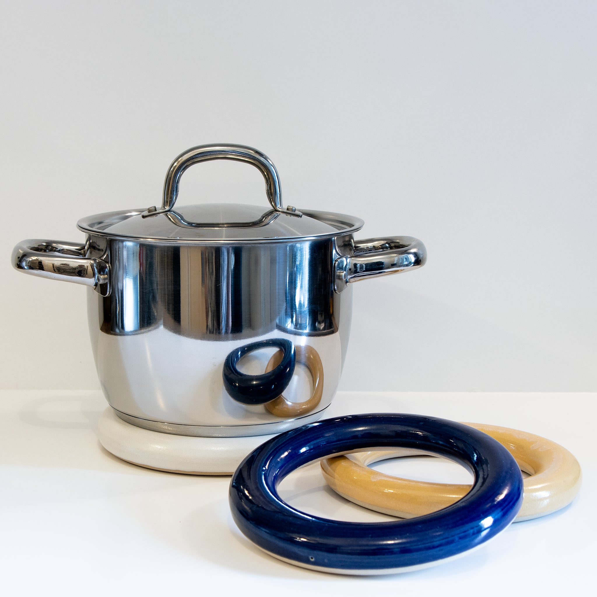 Een glanzende roestvrijstalen pot met deksel rust op een witte onderzetter. Aan de voorkant staan twee ronde keramische onderzetters van AB Ceramics, waaronder de beige en de minimalistische AB Ceramics Pot Onderzetter Geel D16,5, allemaal uitgestald op een wit oppervlak.
