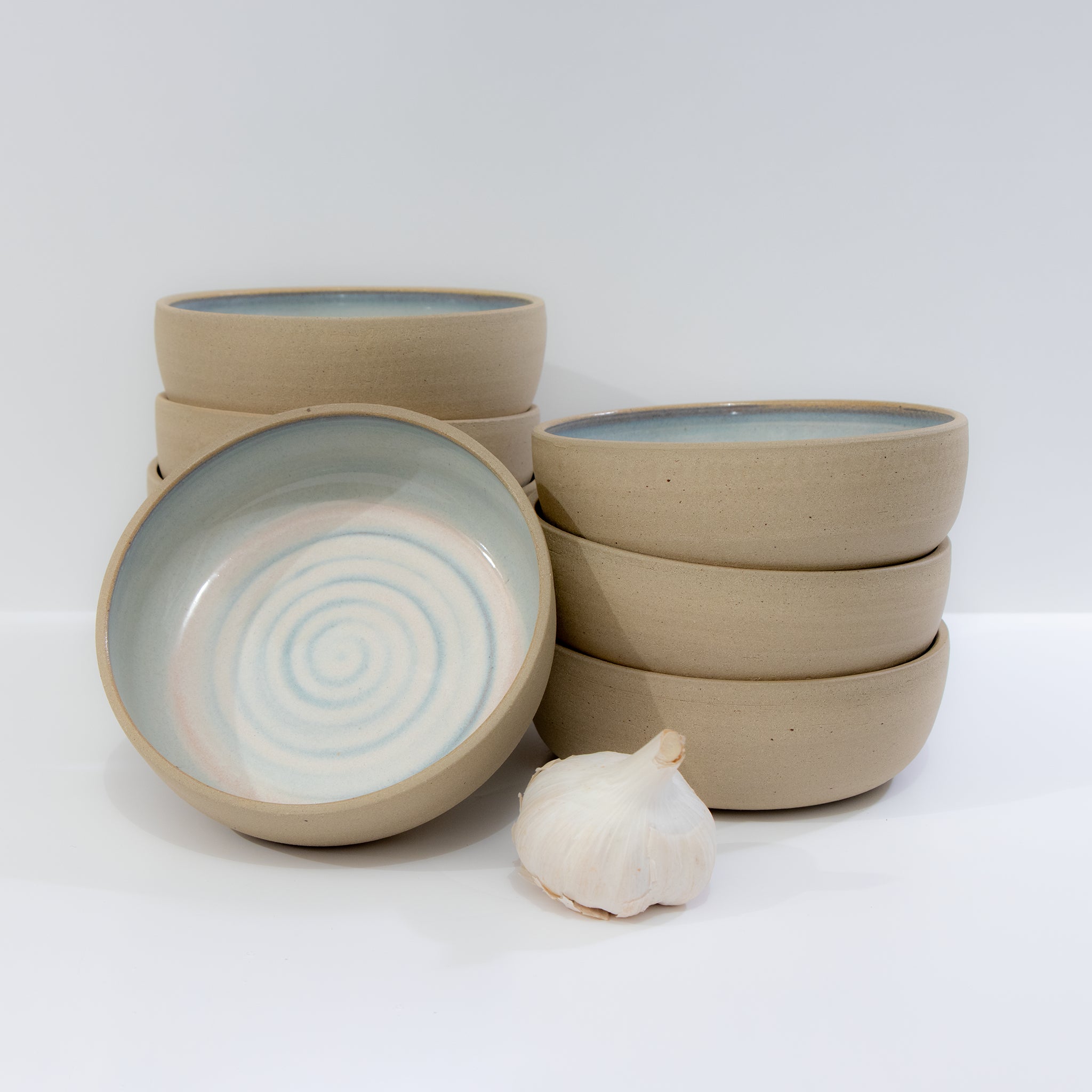 Zes AB Ceramics Schaal Spiraal Geel Opaal D13,5 van steengoed met licht beige buitenkant en geel opaal spiraal binnenkant, handgemaakt, gestapeld naast een knoflookbol tegen een witte achtergrond.