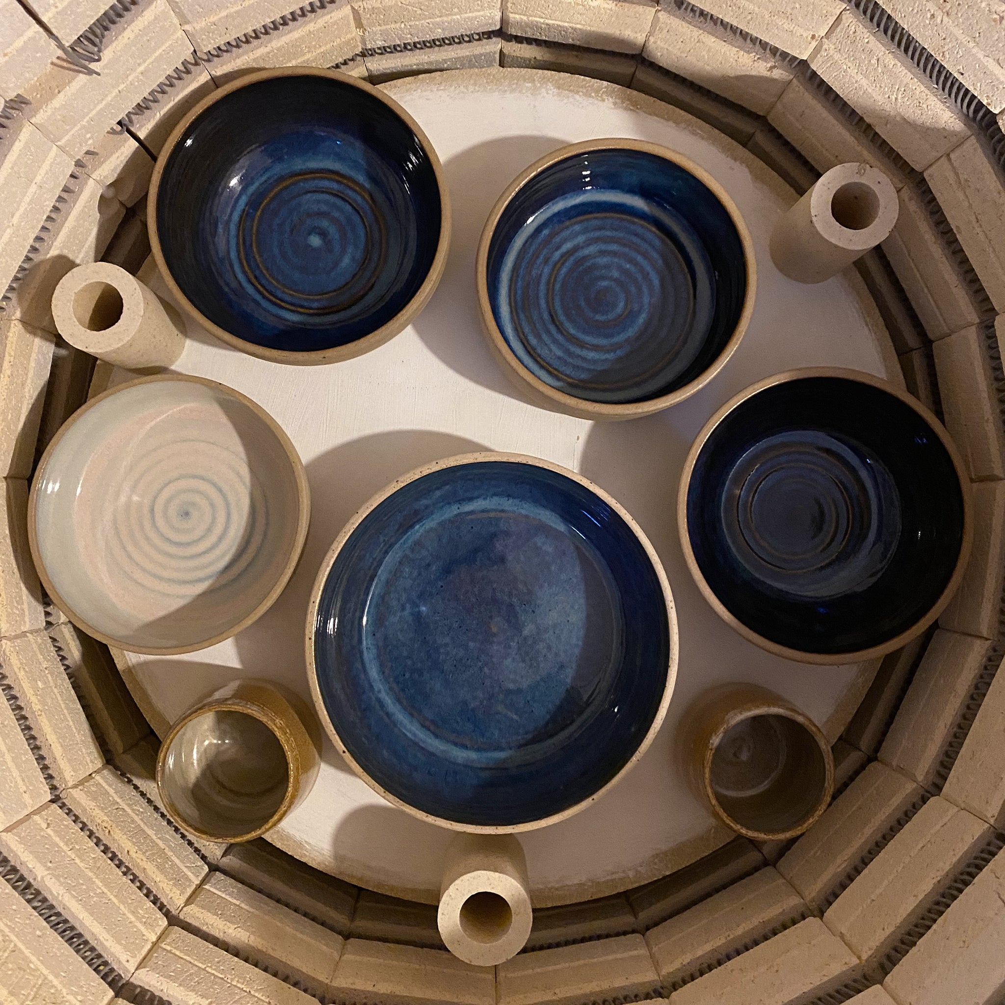 Een bovenaanzicht van zeven AB Ceramics Serveerschaal Spiraal Geel Blauw D21,5 van AB Ceramics, meestal blauw met spiraalmotieven, opgesteld in een cirkel in een oven met beige cilindrische ovenpalen ertussen.