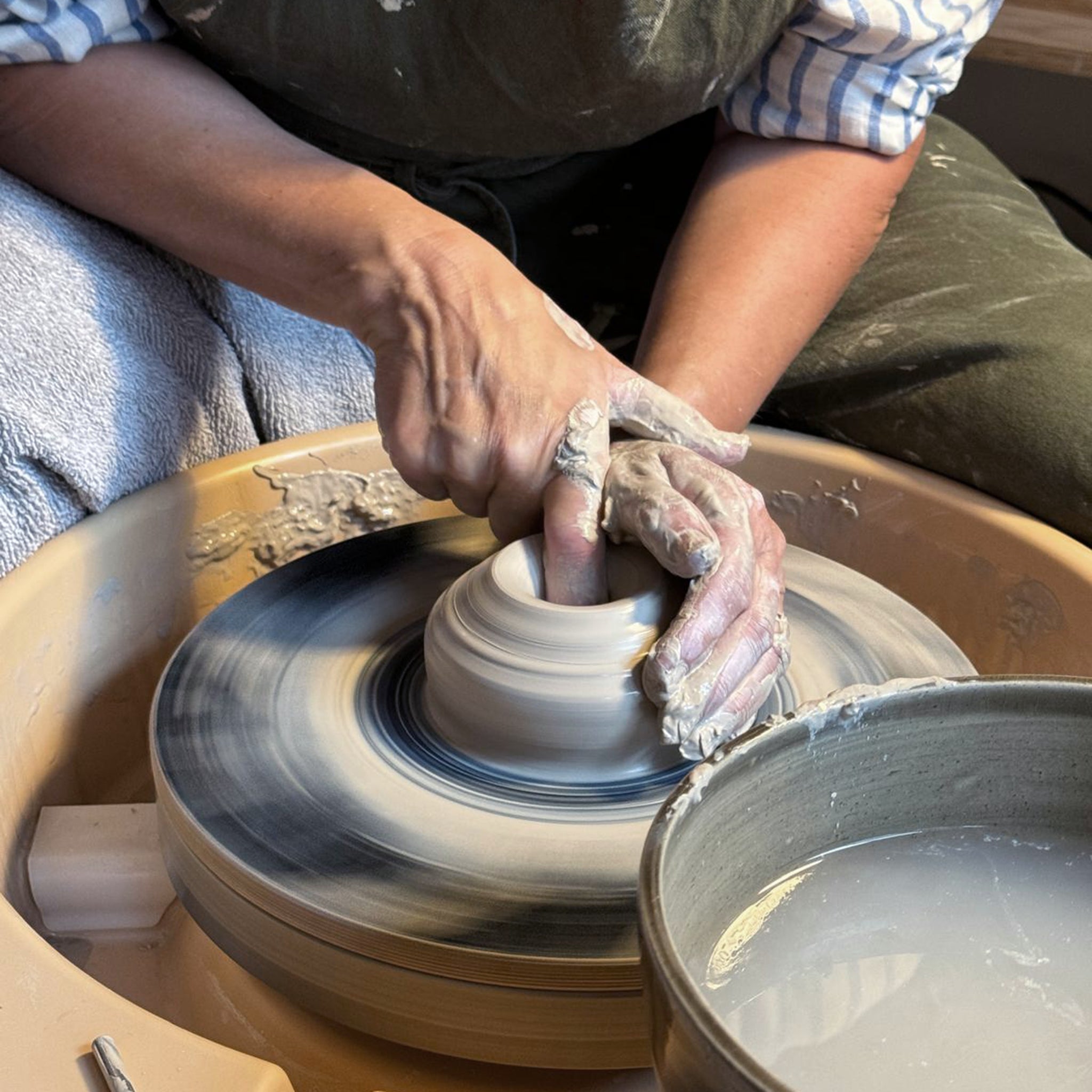 Een kunstenaar vormt steengoed klei op een pottenbakkersschijf, met een geruit overhemd en een schort aan. Kleispetters omringen de AB Ceramics Schaal Spiraal Geel Opaal D13,5 terwijl er een kom water naast staat.
