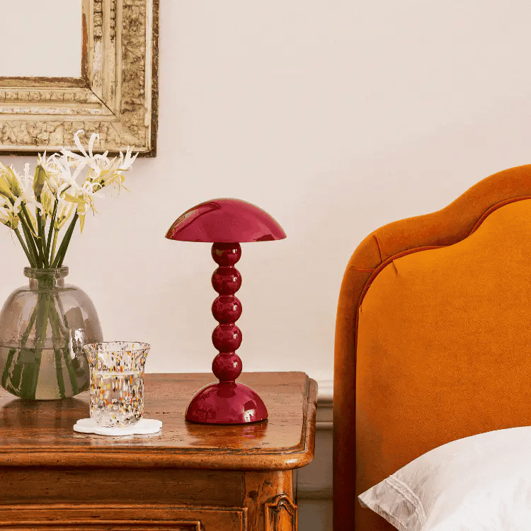 Op een houten nachtkastje staan een vaas met witte bloemen, een figuurglas met een schoteltje en de Addison Ross Bobbin Lamp LED Oplaadbaar Kersenrood; een deel van een oranje gestoffeerd bed en een goudomrande spiegel zijn ook zichtbaar.