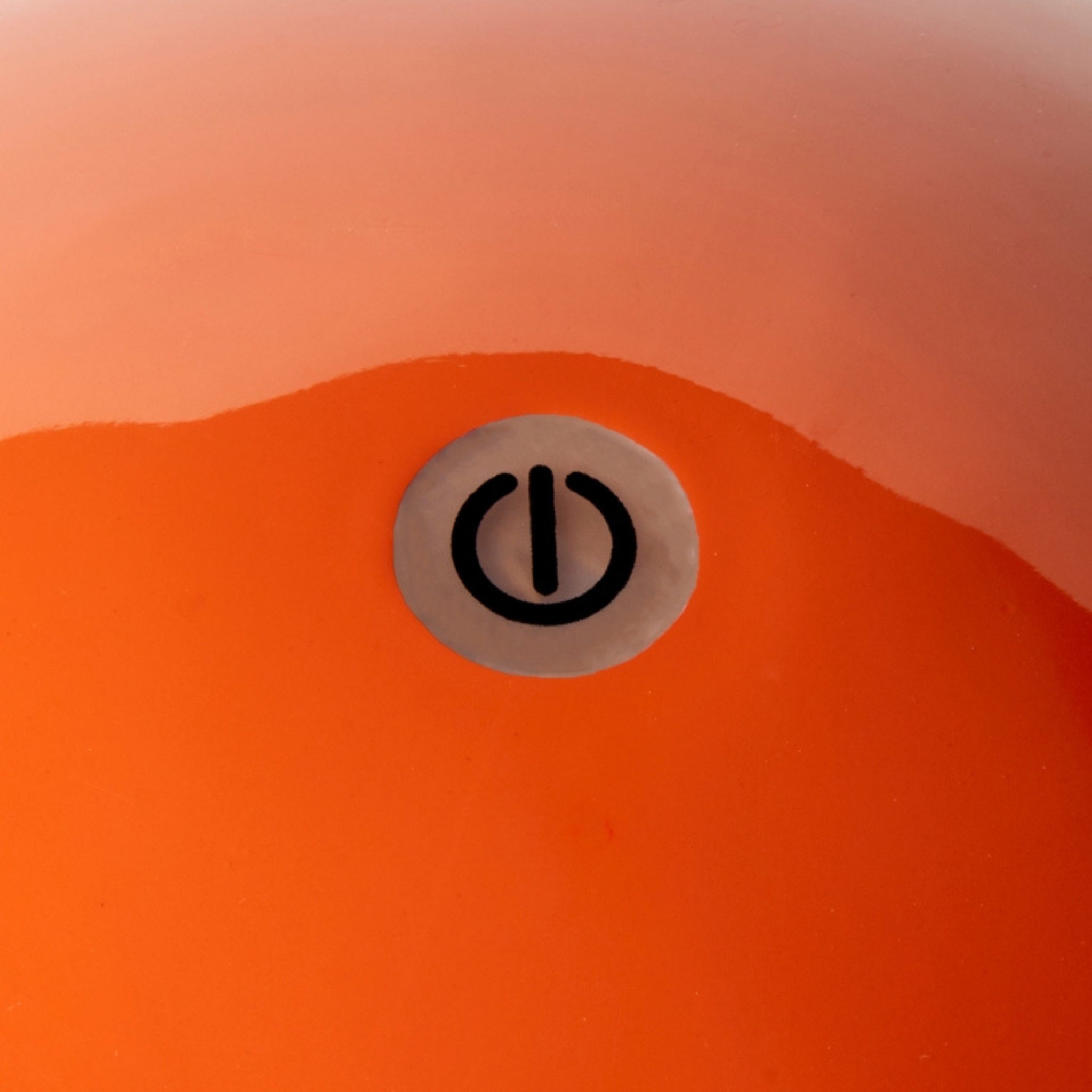 Een close-up van het oppervlak van een oranje lamp met een rond symbool van een aan/uit-knop in het midden, wat het strakke ontwerp oproept van de Addison Ross Bobbin Lamp LED Oplaadbaar Oranje van Addison Ross.
