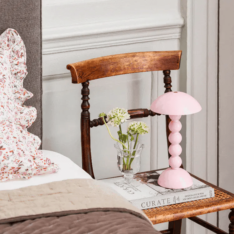 Een houten stoel bij het bed doet dienst als nachtkastje, waarop de Addison Ross Bobbin Lamp LED Oplaadbaar Roze voor sfeerverlichting, een vaas met witte bloemen en boeken staan. Het bed is opgemaakt met een gebloemd kussen en een knus beige dekbed.