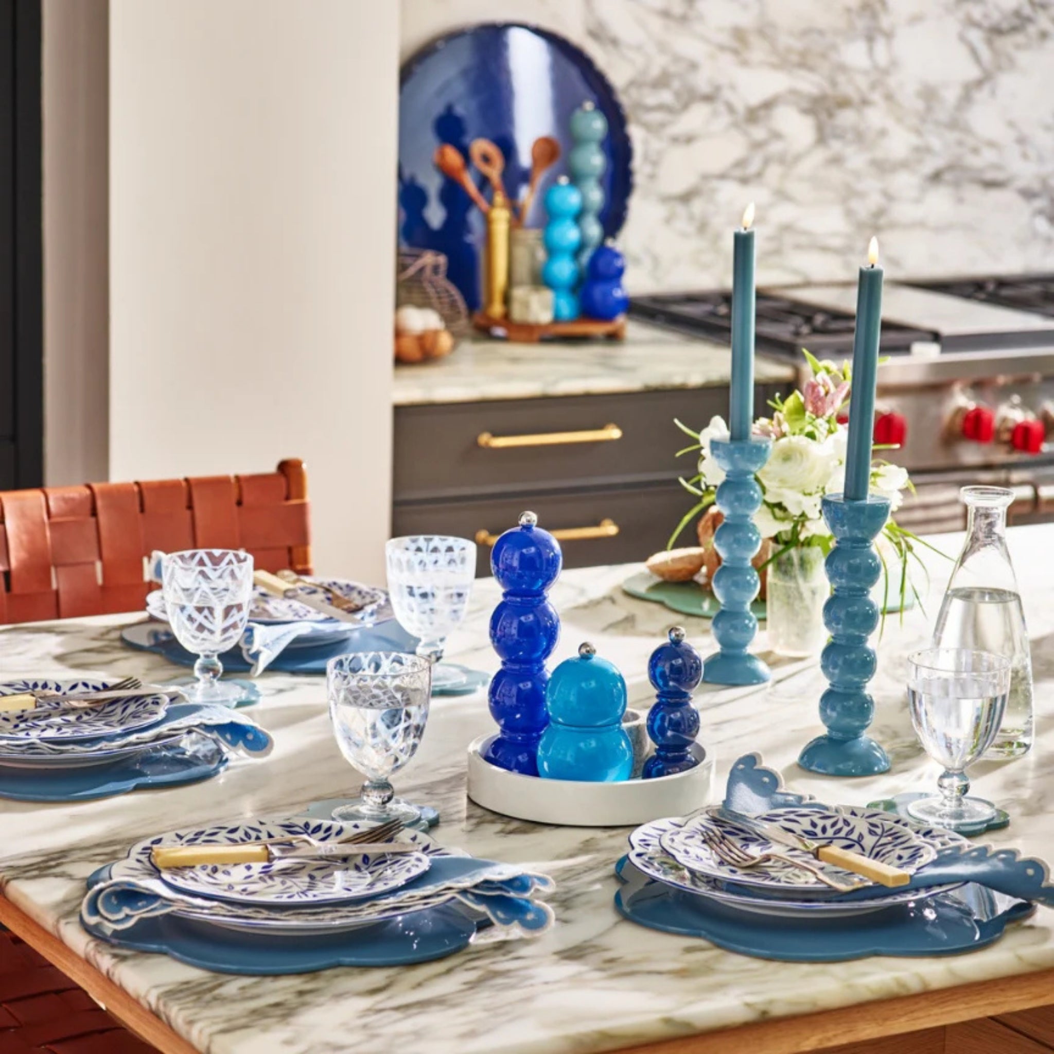 Op een marmeren keukentafel voor vier personen staan blauwe en witte borden, kobaltkleurig glaswerk, blauwe kandelaars met hoge kaarsen, een middenstuk van gestapelde blauwe glazen bollen, een Addison Ross Bobbin Zout-en Pepermolen Kobalt 24 cm en verse bloemen.