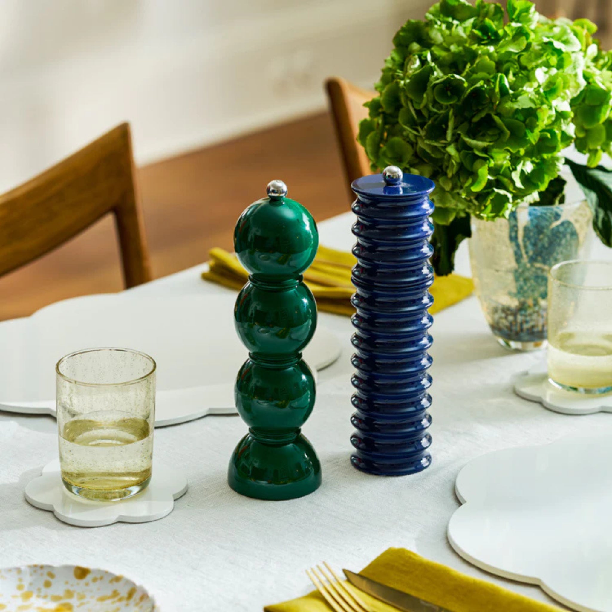 Een gedekte eettafel met borden, glazen, gele servetten, een groene ronde Addison Ross Bobbin zout-en pepermolen in Racing Groen (24 cm), een hoge blauwe molen en een boeket groene hortensia's.