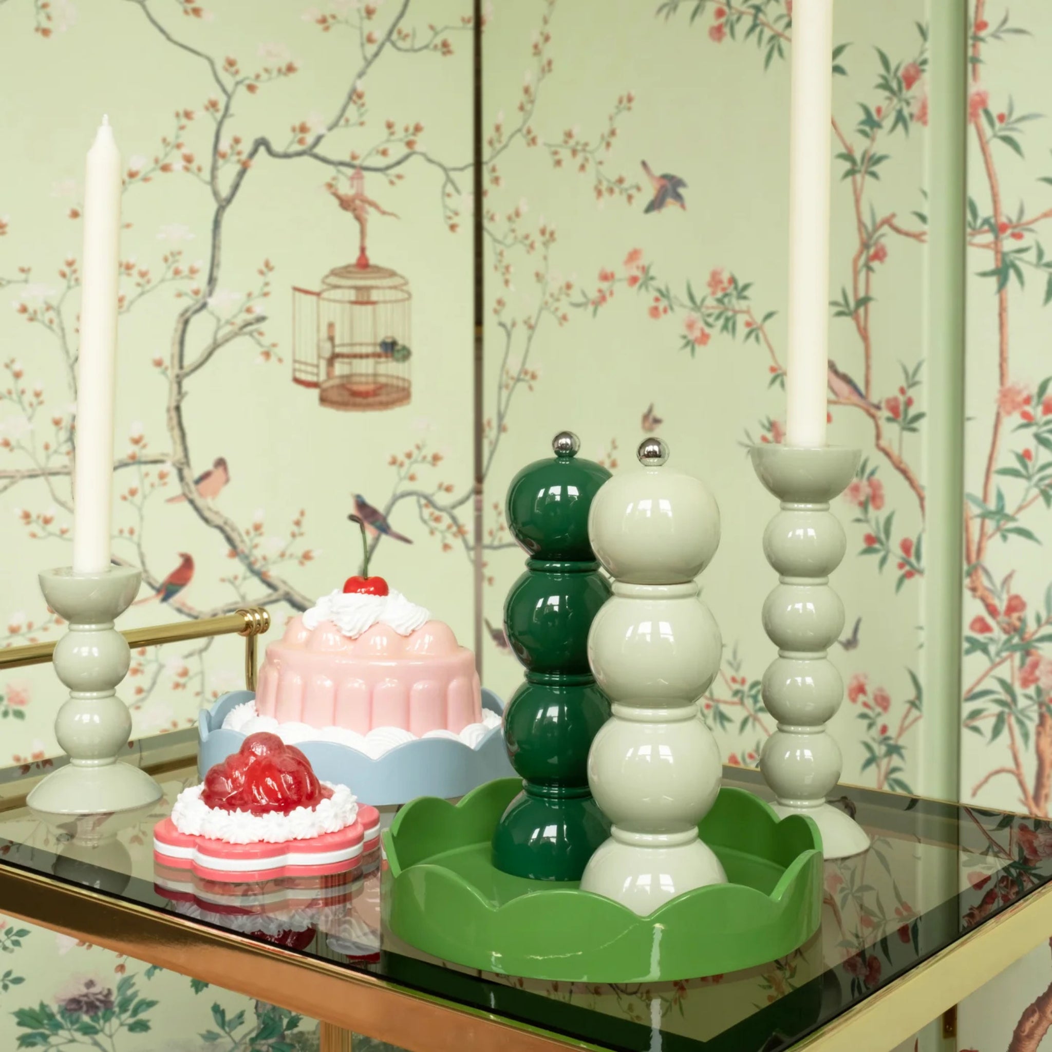 Een decoratieve tafel met Addison Ross Bobbin kandelaars met witte kaarsen, een groene Addison Ross Bobbin Zout-en Pepermolen Racing Groen 24 cm, twee desserts, alles tegen vogel- en takkenbehang met een vogelkooi.