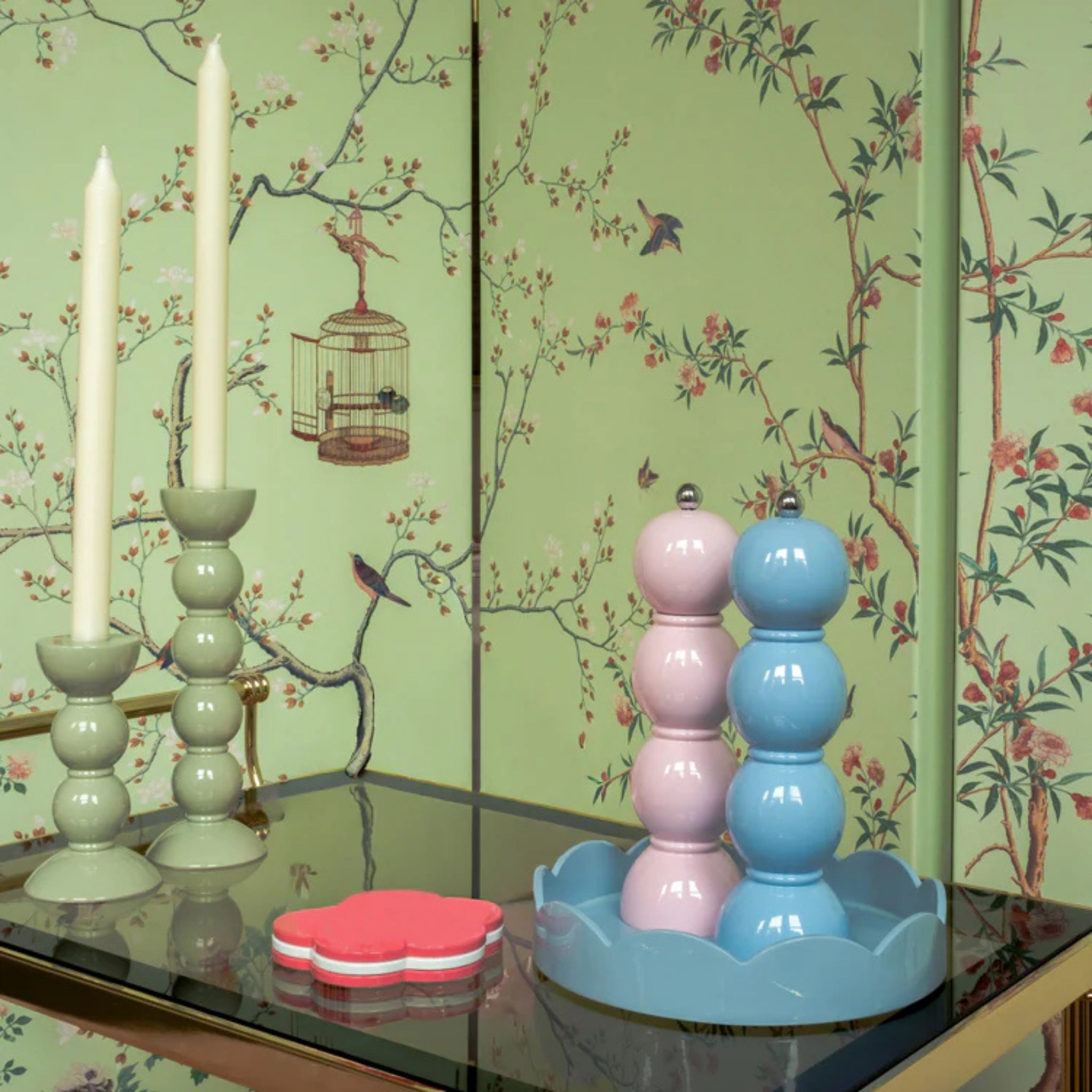 Een pastelkleurige tafelschikking met twee hoge witte kaarsen in lichtgroene houders, een roze bloemenonderzetter en de Addison Ross Bobbin Zout-en Pepermolen Roze 24 cm met keramisch maalwerk op een blauw dienblad tegen groen vogel- en takbehang.