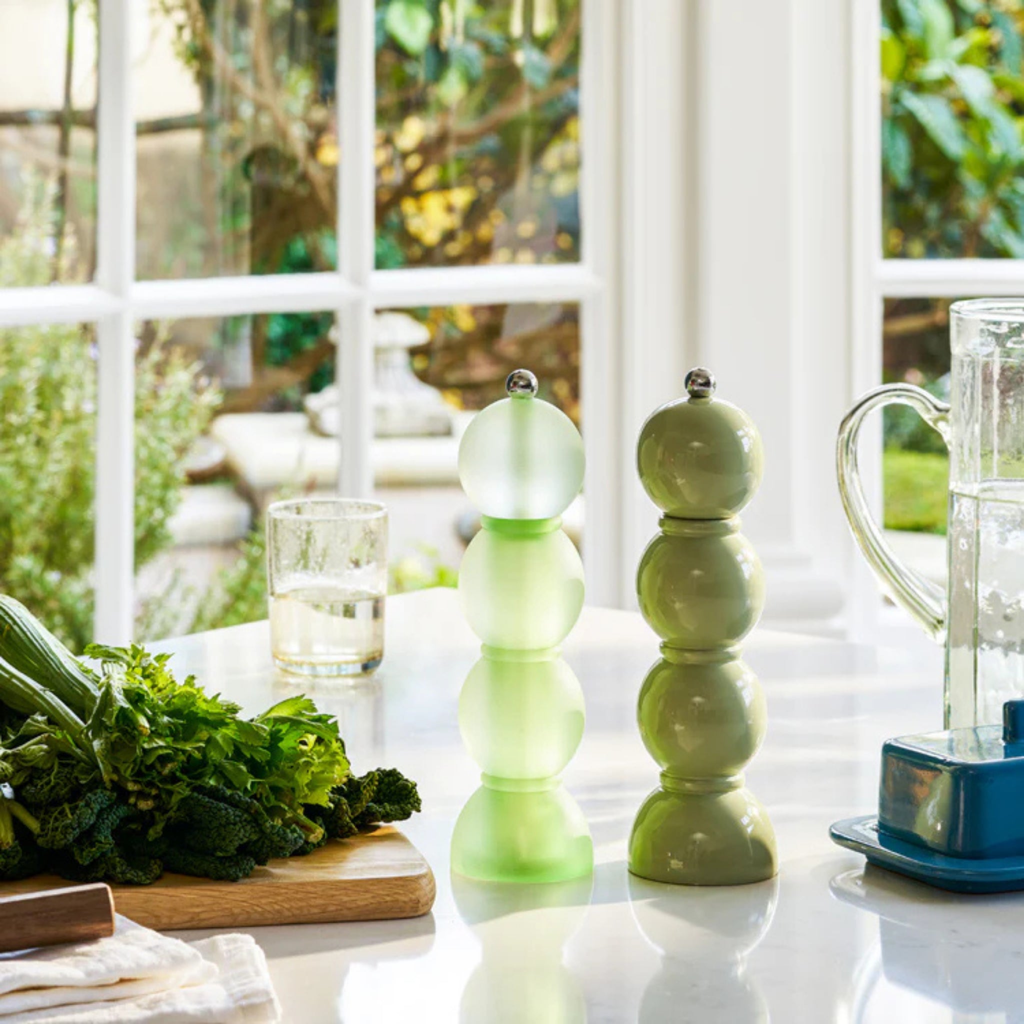 Op een zonovergoten aanrecht staan twee Addison Ross Bobbin zout-en pepermolens in saliegroen (24 cm), een snijplank met bladgroen, een glas water en een kan bij grote ramen met uitzicht op een tuin.
