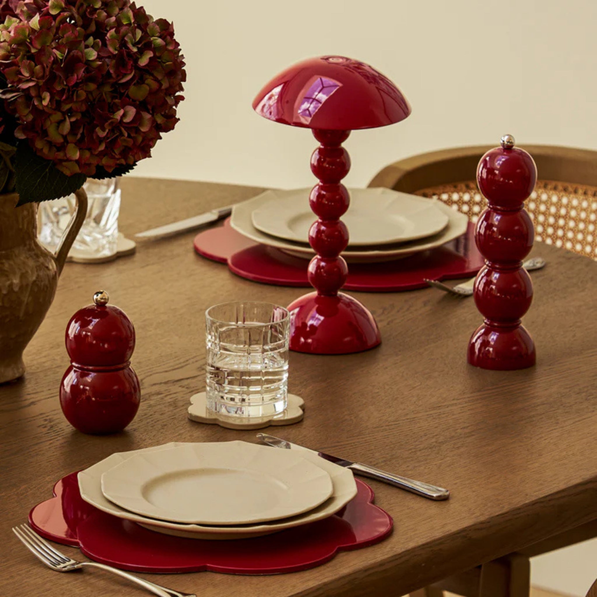 Een houten gedekte eettafel met crèmekleurige borden op rode placemats, heldere glazen, een Addison Ross Bobbin Lamp LED Oplaadbaar Kersenrood, bijpassend rood peper- en zoutstel en een vaas met hortensia's.