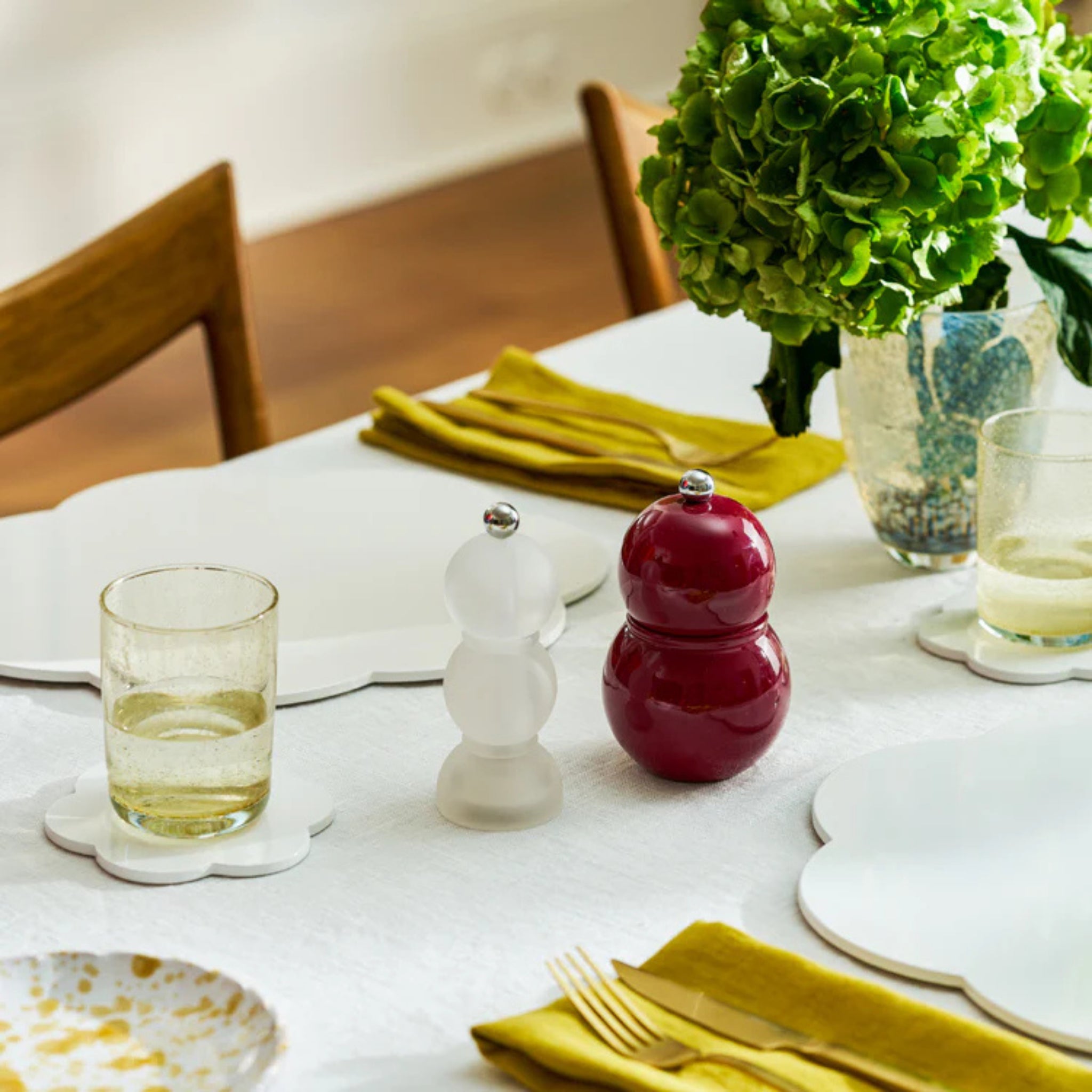 Een gedekte eettafel met gele servetten, borden, glazen water, een Addison Ross Chubby Zout-en Pepermolen Kersenrood 12 cm, en een vaas met groene hortensia's, met houten stoelen op de achtergrond.