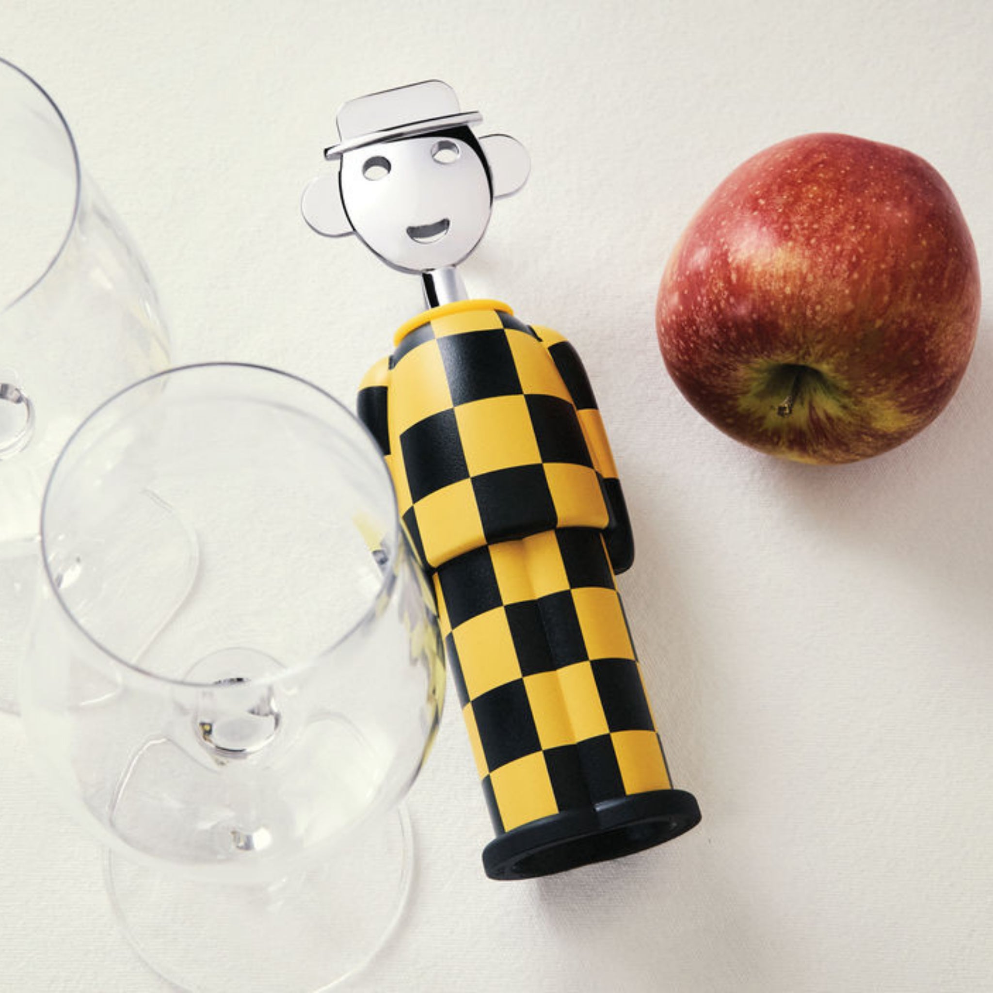 De Alessi Alessandro Kurkentrekker Giallo e Nero flesopener, met Italiaans geïnspireerd zwart-geel geruit ontwerp, rust op een wit oppervlak naast een appel en twee lege wijnglazen.