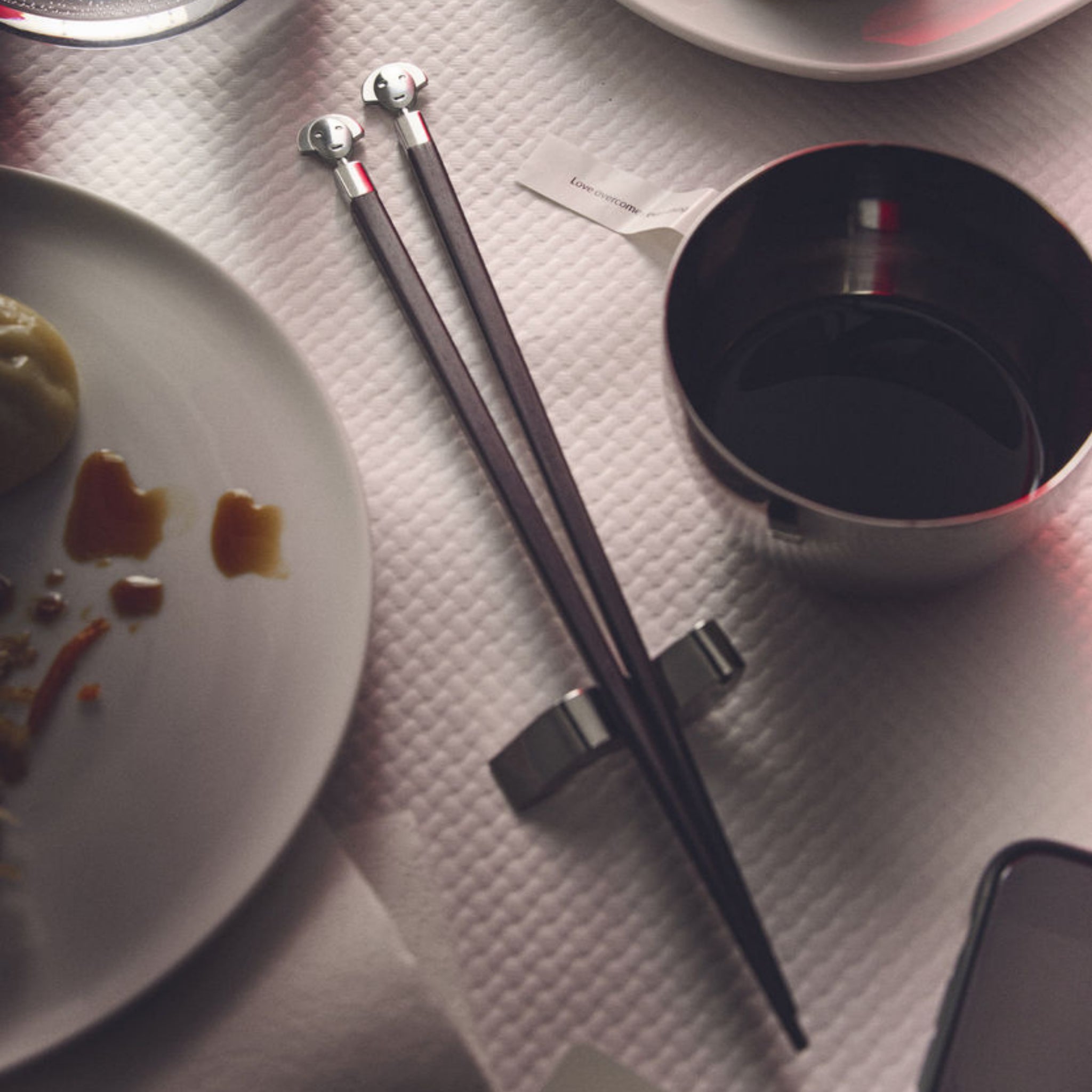 Een paar Alessi Anna eetstokjes + Houder van Alessi liggen naast een kopje donkere vloeistof, een wit bord met etensresten en een wit tafelkleed met structuur.