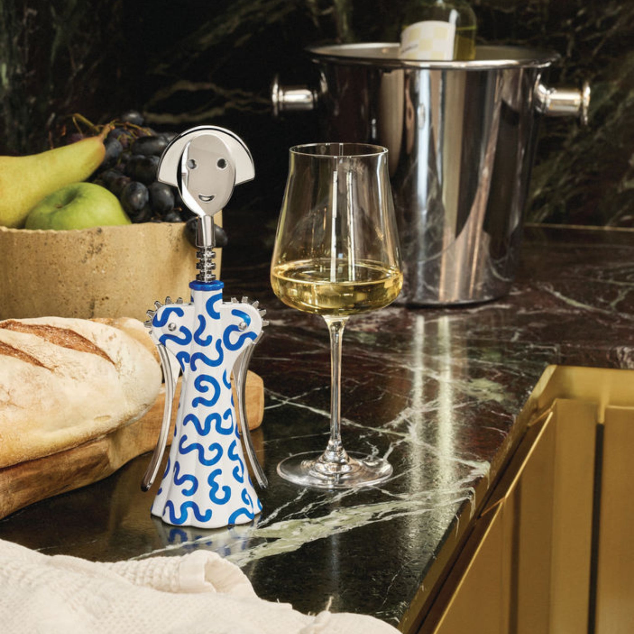 Een Alessi Anna G Kurkentrekker Delft kurkentrekker, een glas witte wijn, brood, fruit en een wijnemmer met een fles staan opgesteld op een donker marmeren aanrecht.
