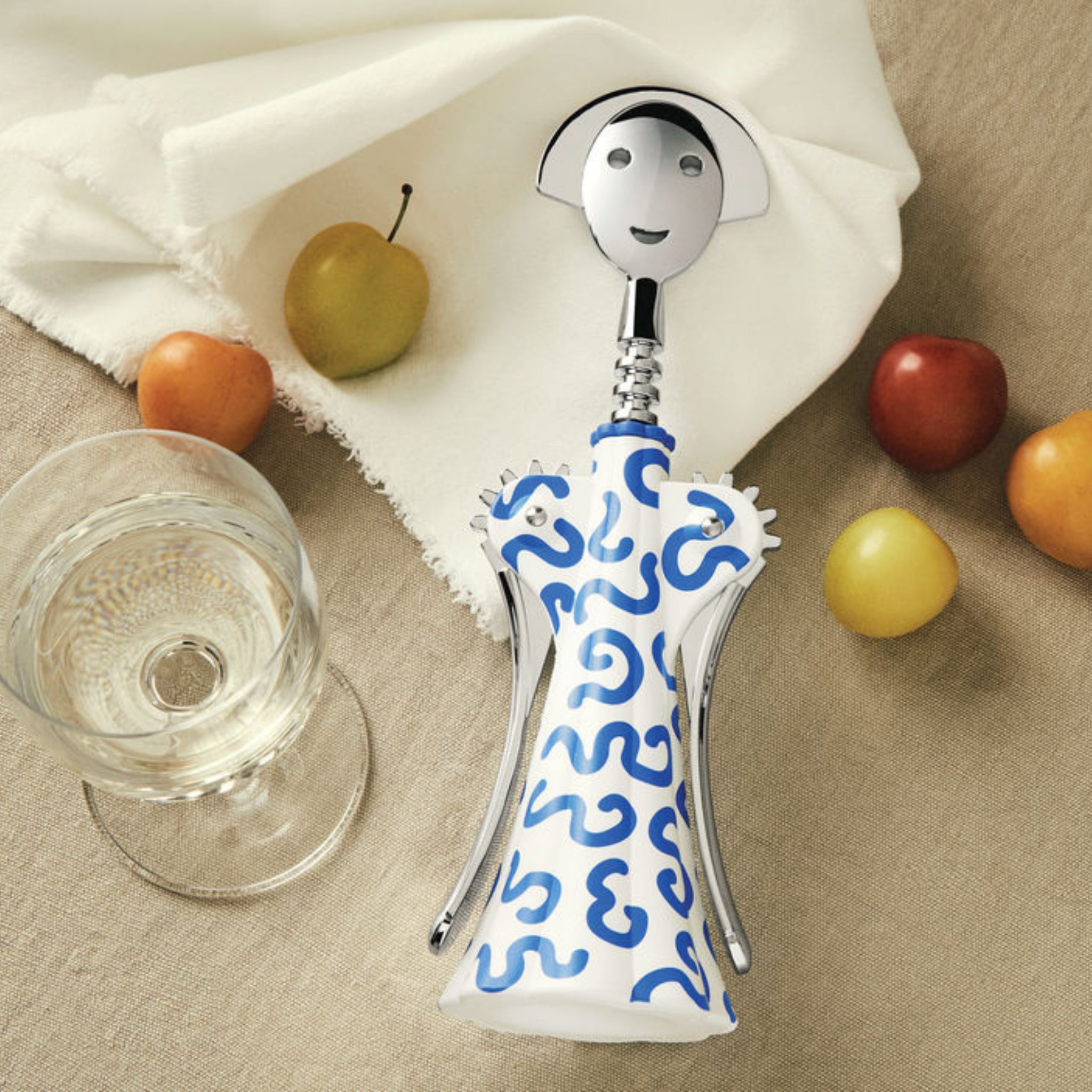 De Alessi Anna G Kurkentrekker Delft van Alessi, met een lachend gezichtje en Delftsblauw ontwerp, rust op een beige tafelkleed naast een glas witte wijn, een wit servet en verschillende kleurrijke vruchten.