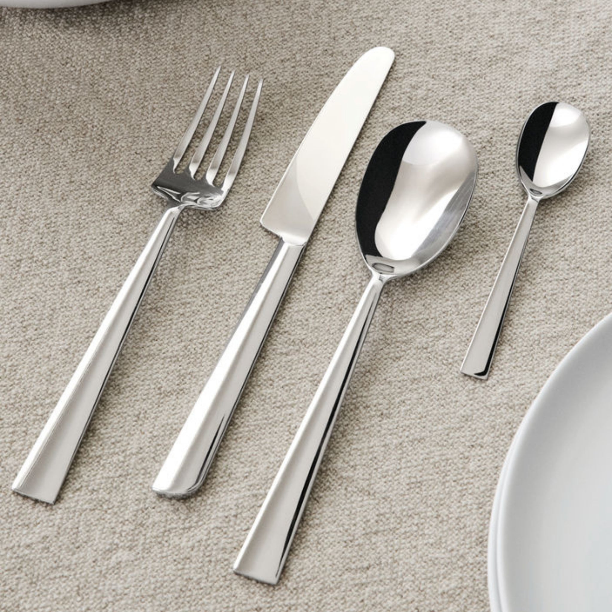 Vier glanzend zilveren bestekken uit de Alessi Asta Bestek 24-delig set van Alessi-een vork, mes, eetlepel en theelepel-zijn netjes geplaatst op een beige tafelkleed met textuur naast een wit bord.