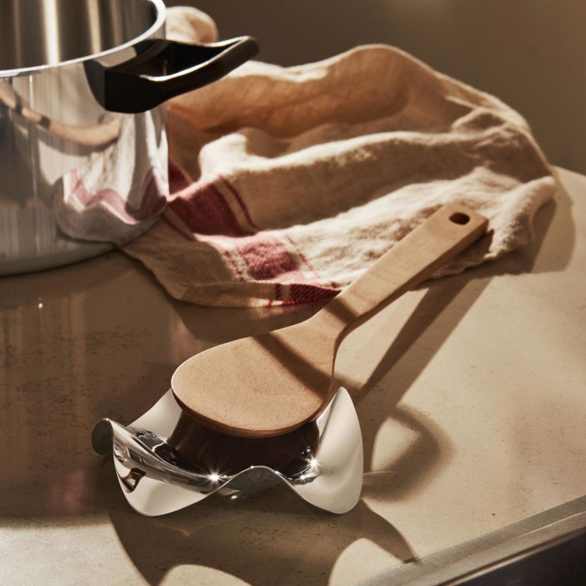 Een houten lepel rust op een Alessi Blip Lepelhouder, een stijlvol Italiaans ontworpen keukenaccessoire van Alessi, naast een pan en een beige handdoek met een rode streep op het aanrecht.