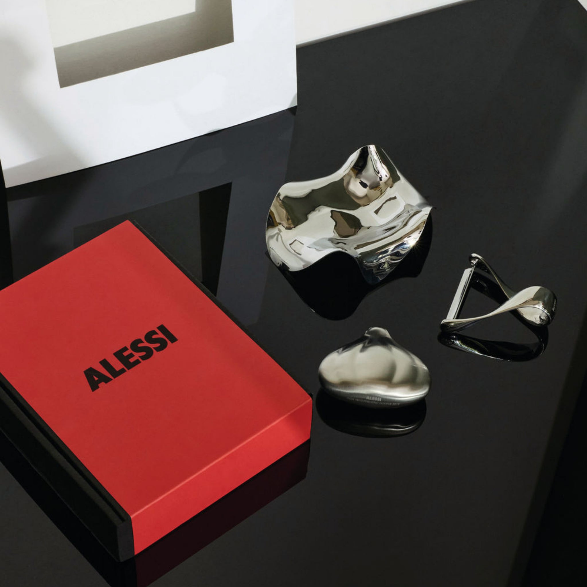 Een glanzend zwarte tafel met een rood en zwart Alessi-doosje, een glanzend abstract zilveren schaaltje, een Alessi Blip Lepelhouder van Alessi en een zilveren keukenaccessoire in de vorm van knoflook. Een wit geometrisch object op de achtergrond accentueert het Italiaanse design.