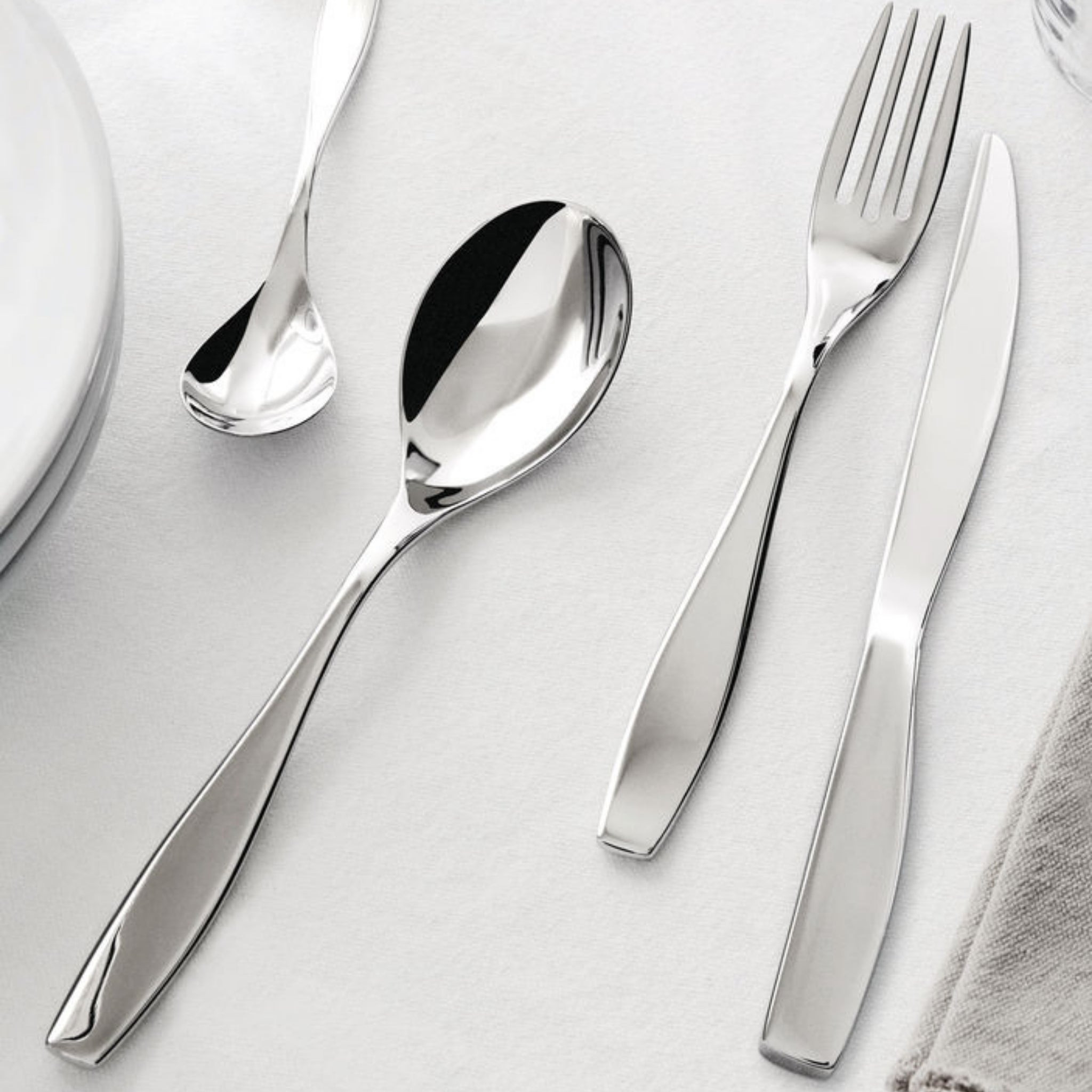 Een Alessi Duna Bestek 24-delig servies met Italiaans design staat op een wit tafelkleed naast een bord en servet.