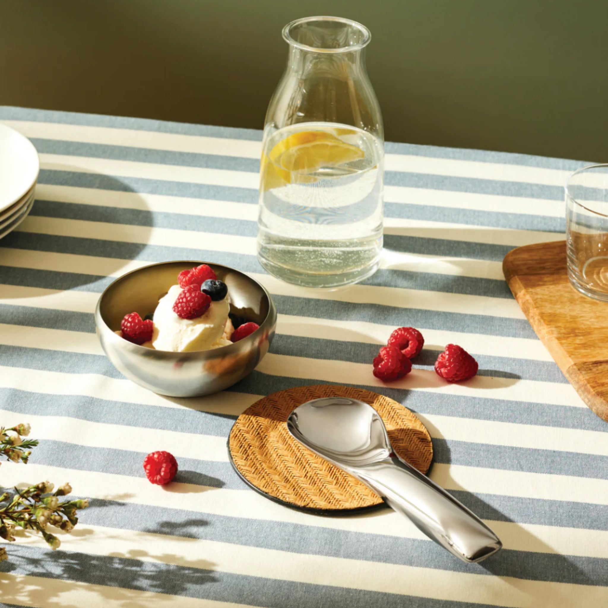 Een gestreept tafelkleed toont een kom ijs met bessen, een karaf citroenwater, een glas en een Alessi Koki Ijslepel op een onderzetter, met frambozen verspreid.