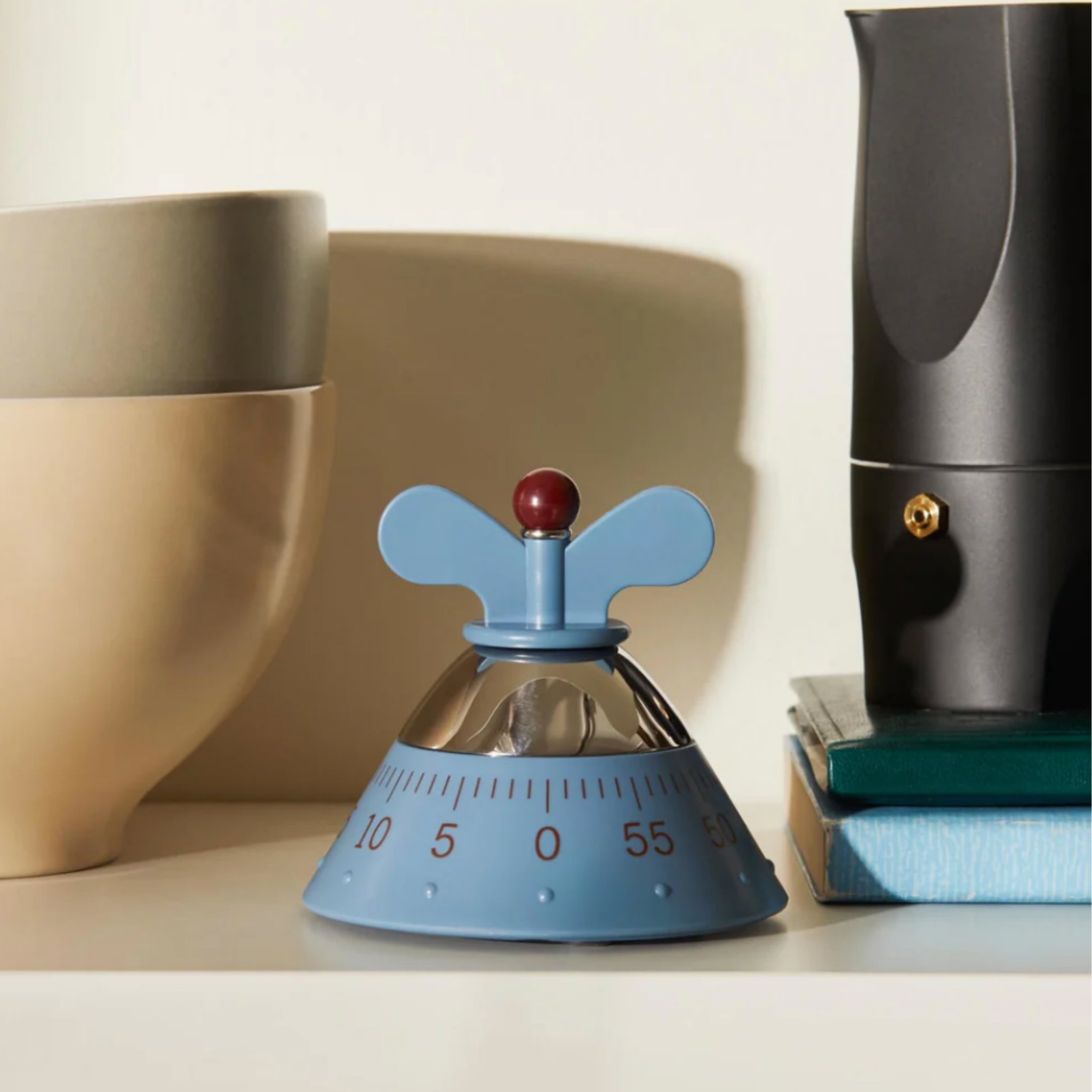 De Alessi Kookwekker Blauw, een ronde blauwe keukenwekker met een rode knop en vleugelvormige handgrepen, staat op een plank naast gestapelde keramische kommen, twee boeken en een zwarte koffiepot - een opvallende keukenaccessoire.