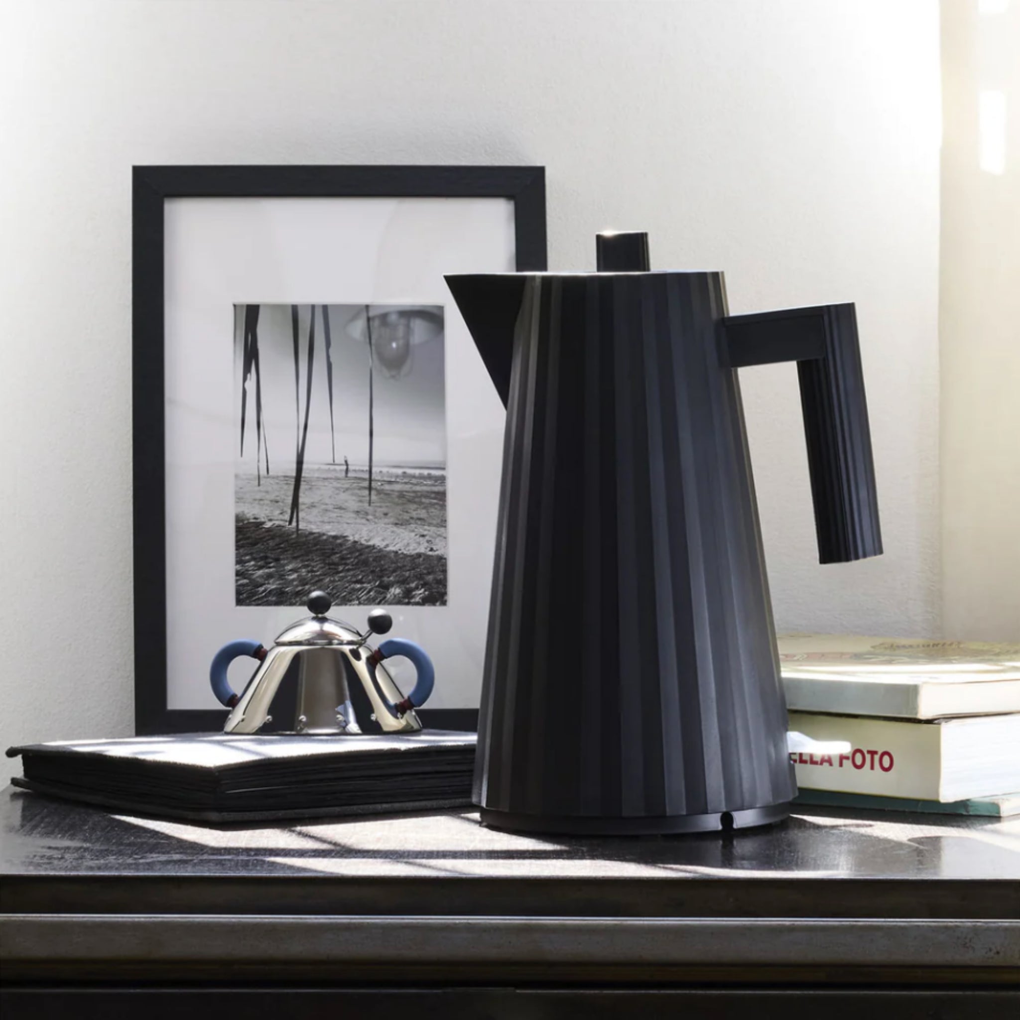 Een moderne zwarte koffiepot staat op een donkere tafel naast een ingelijste zwart-wit strandfoto, een Alessi Plissé Elektrische Waterkoker Zwart 1,7 l, een stapel zwarte servetten en een gedeeltelijk zichtbaar boek met de titel FOTO.