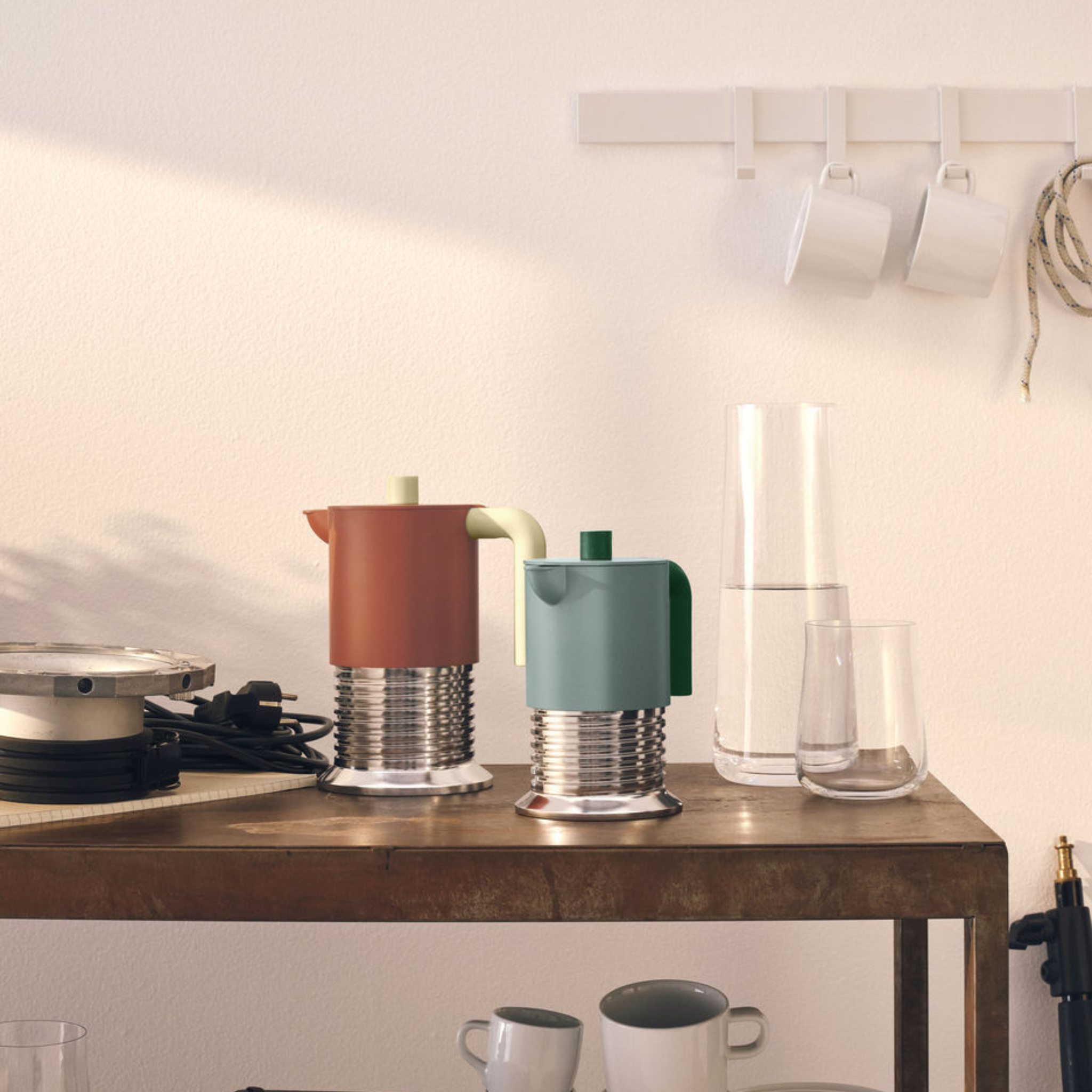 De Alessi Vite Koffiemaker Saliegroen 3-Kops van Alessi staat op een rustieke houten tafel met mokken, glazen en een karaf. Drie witte kopjes hangen aan een nabijgelegen muur. Perfect voor inductiegebruik en stijlvolle koffiemomenten.