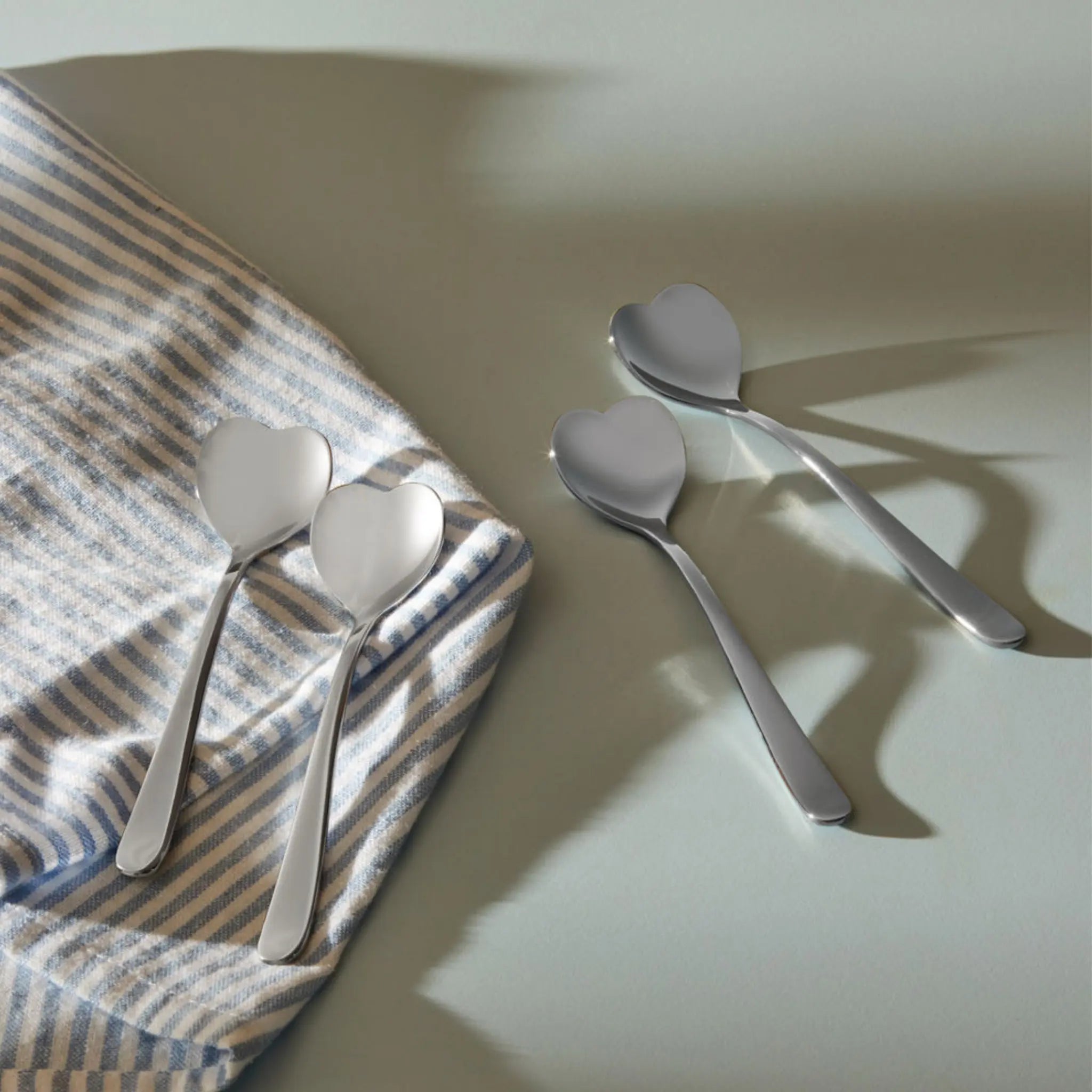 Vier Alessi Big Love theelepels (set van 4), ontworpen door Miriam Mirri, liggen op een lichte ondergrond: twee op een blauw-wit gestreepte doek en twee ernaast. De zilverkleurige lepeltjes werpen zachte schaduwen.