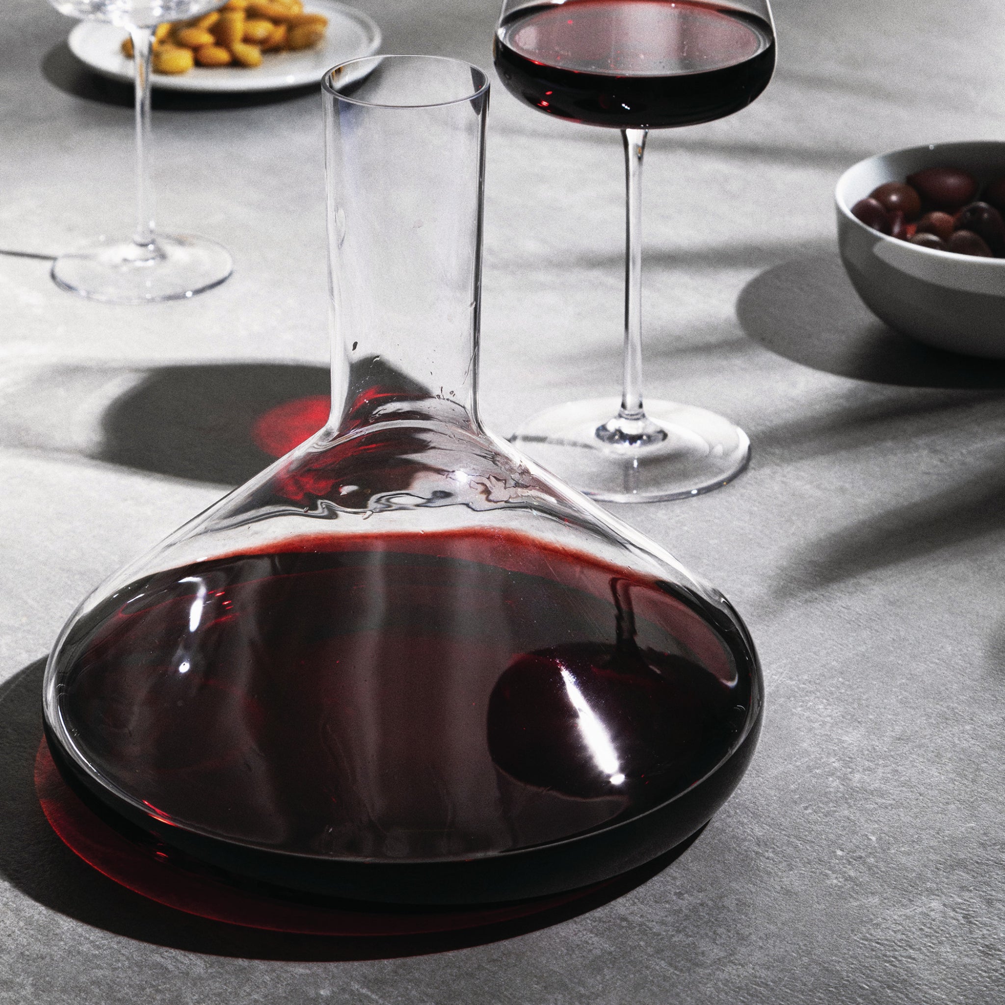 Een Alessi Eugenia Decanteerkaraf 75 cl, gevuld met rode wijn, rust op een grijze tafel naast een wijnglas, olijven en hapjes. Licht en schaduw weerkaatsen op het kristallijne glas van Alessi voor een elegante touch.