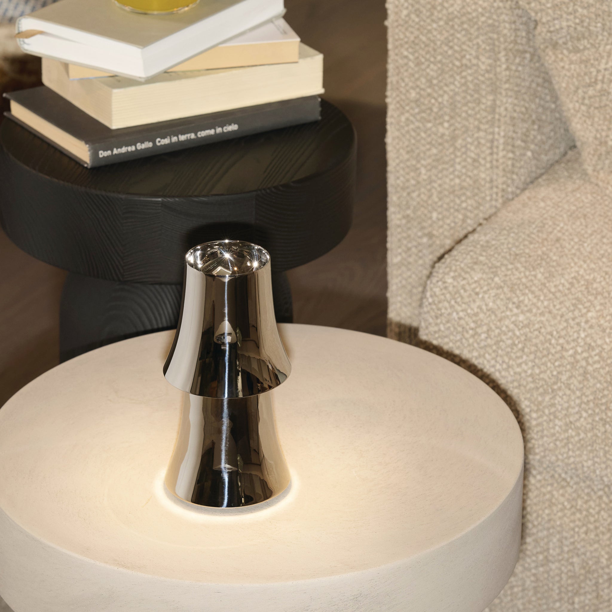 De Alessi Halesia Tafellamp LED Oplaadbaar staat op een ronde, lichte bijzettafel naast een beige bank met textuur, terwijl gestapelde boeken op een zwart tafeltje op de achtergrond rusten en een gezellige en eigentijdse sfeer creëren.