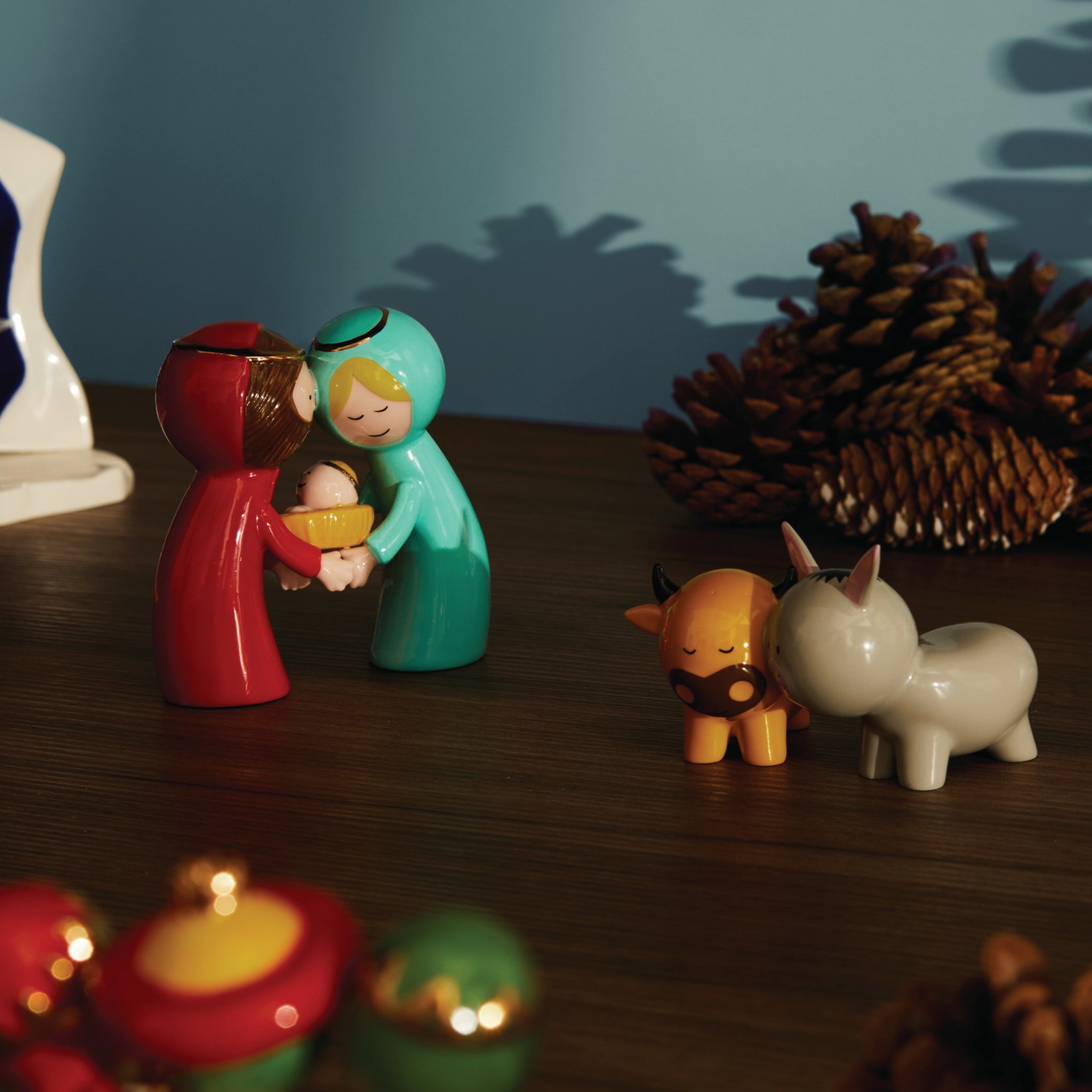 Twee levendige Alessi Happy Eternity babyfiguren tonen Maria en Jozef met baby Jezus in een kerststal, omringd door een ezel, os, dennenappels en kerstornamenten op een warm verlicht houten oppervlak.