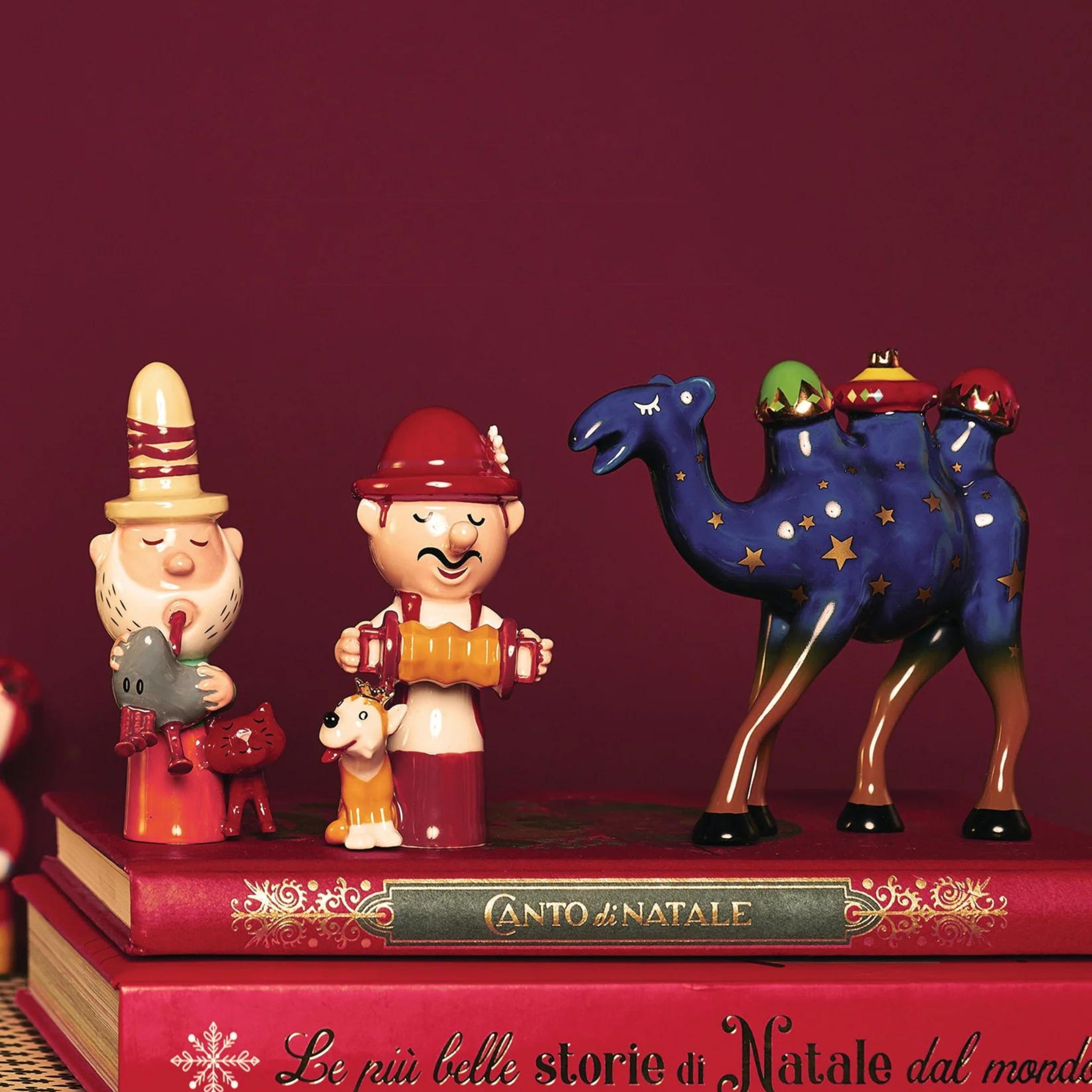 De Alessi Happy Eternity Baby Eolo De Doedelzakspeler van Alessi staat tussen levendige porseleinen kerstfiguren op rode boeken, tegen een bordeauxrode achtergrond, die de speelse geest van de feestdagen vastlegt.