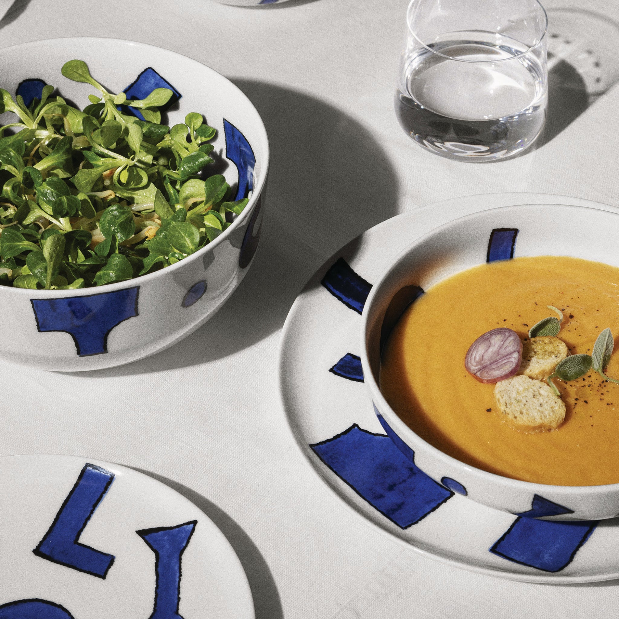 Een frisse bladsalade in de Alessi Itsumo Saladekom Wit 195 cl, naast sinaasappelsoep met croutons, kruiden en radijs in Japans geïnspireerde blauwe en witte borden op een wit tafelkleed, staat naast een helder glas water.