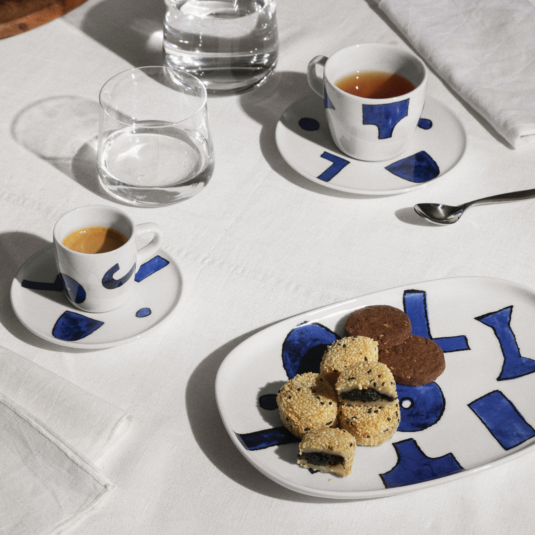 Een gedekte tafel met Alessi Itsumo Koffiekop + Schotel Wit 21 cl, thee of koffie, water, koekjes en gebakjes op een bord en een lepel - allemaal netjes gerangschikt op een wit tafelkleed met een servet in verfijnd Japans design.
