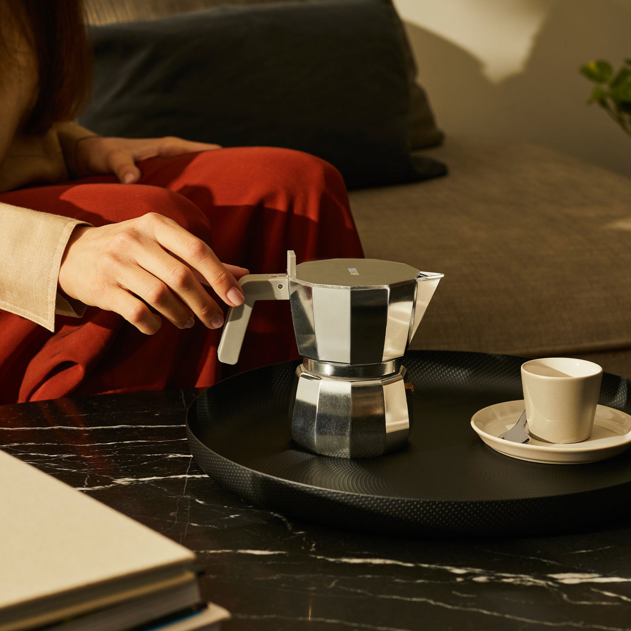 Een persoon in een rode broek zit op een bank en reikt naar een Alessi Moka Espresso Koffiemaker 3-kops op een zwart dienblad met een klein kopje en schoteltje, op een donker marmeren tafel en badend in warm, op Chipperfield geïnspireerd zonlicht.