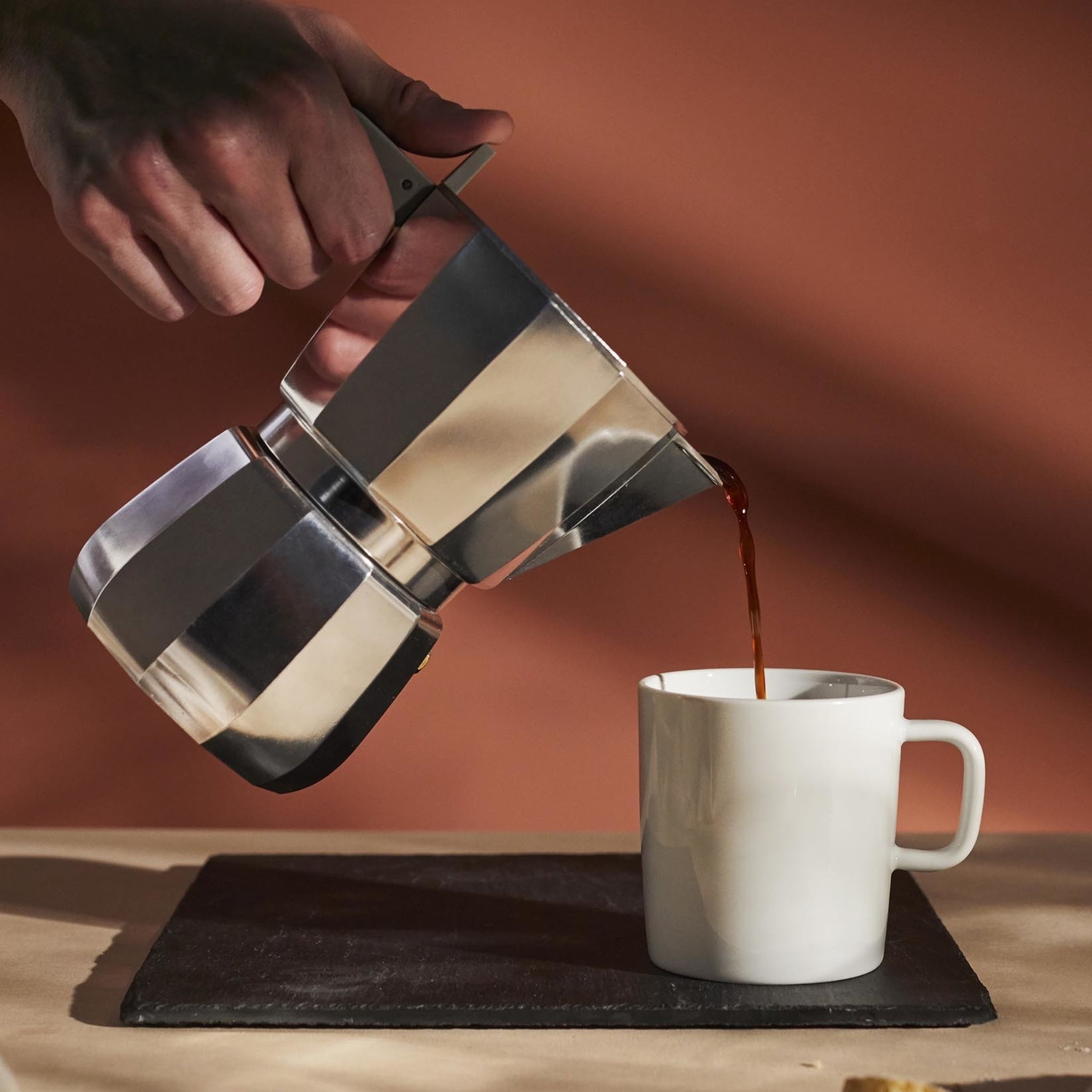 Een hand schenkt koffie uit de Alessi Moka Espresso Koffiemaker 6-kops in een witte keramische mok op een donker dienblad, met een warmbruine achtergrond en zachte verlichting.