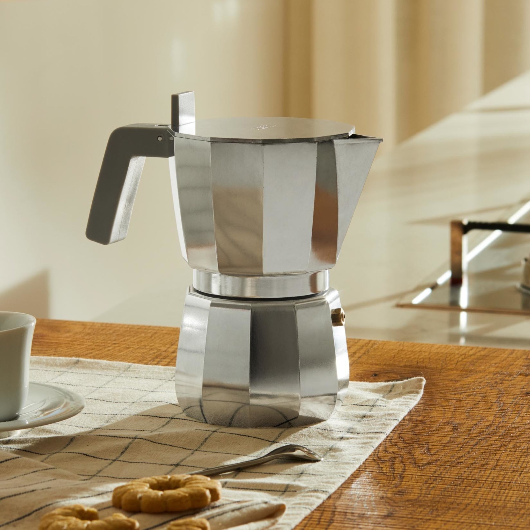 De Alessi Moka Espresso Koffiemaker 9-kops van Alessi ligt op een geruit kleed gedrapeerd over een houten tafel, vergezeld van een kop, schotel, lepel en koekjes. Zonlicht vult de keuken en roept een warme en uitnodigende sfeer op.