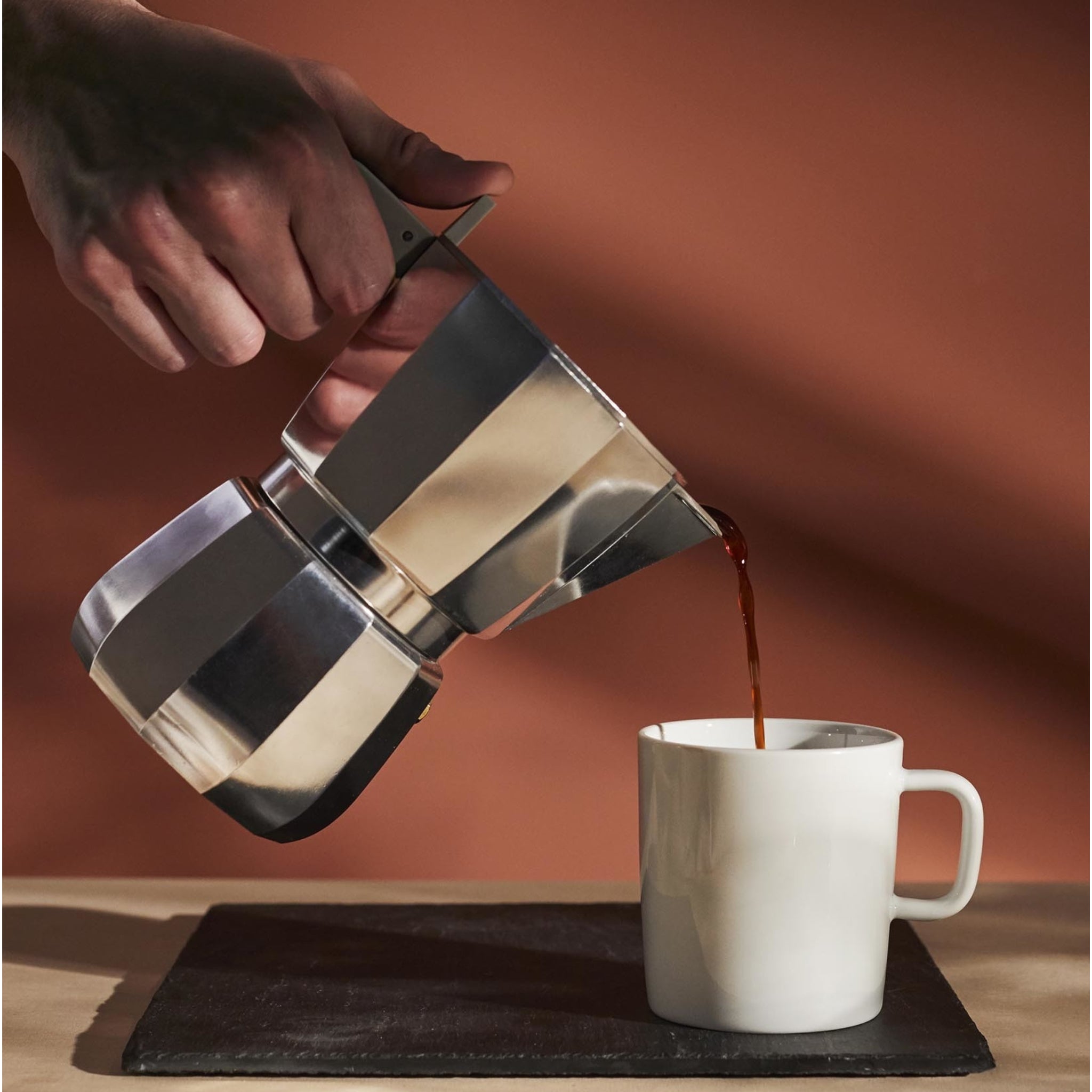 Een hand schenkt koffie uit de Alessi Moka Espresso Koffiemaker 9-kops in een witte keramische mok op een zwarte mat, tegen een warmbruine achtergrond, die de minimalistische esthetiek van Alessi weergeeft.