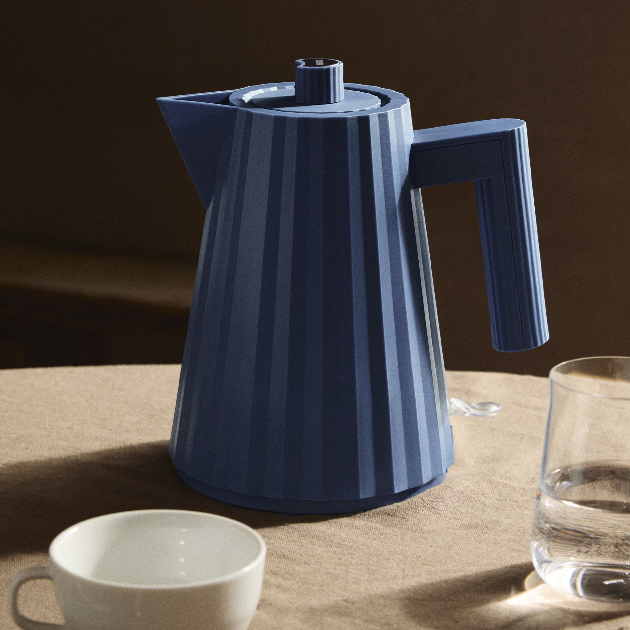 De Alessi Plissé Elektrische Waterkoker Blauw 1 l, met zijn moderne geribbelde ontwerp, staat op een beige tafelkleed naast een witte beker en glas water in warm, zacht licht.