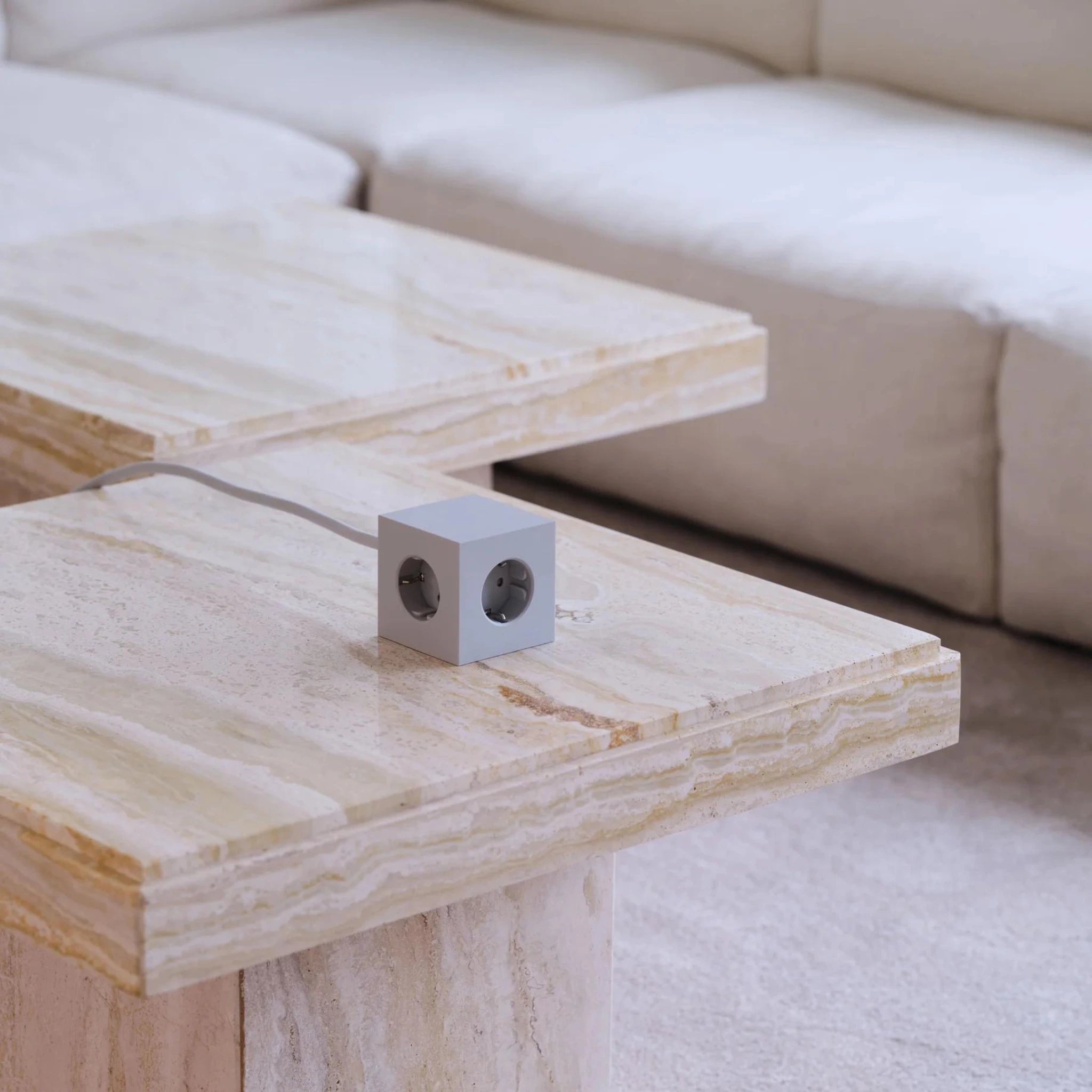 Een Avolt Square 1 USB-C Stekkerdoos Gotland Grijs 1,8 m stekkerdoos met twee zichtbare stopcontacten is geplaatst op een licht marmeren salontafel voor een beige bank en tapijt, met handige stopcontacten met USB.