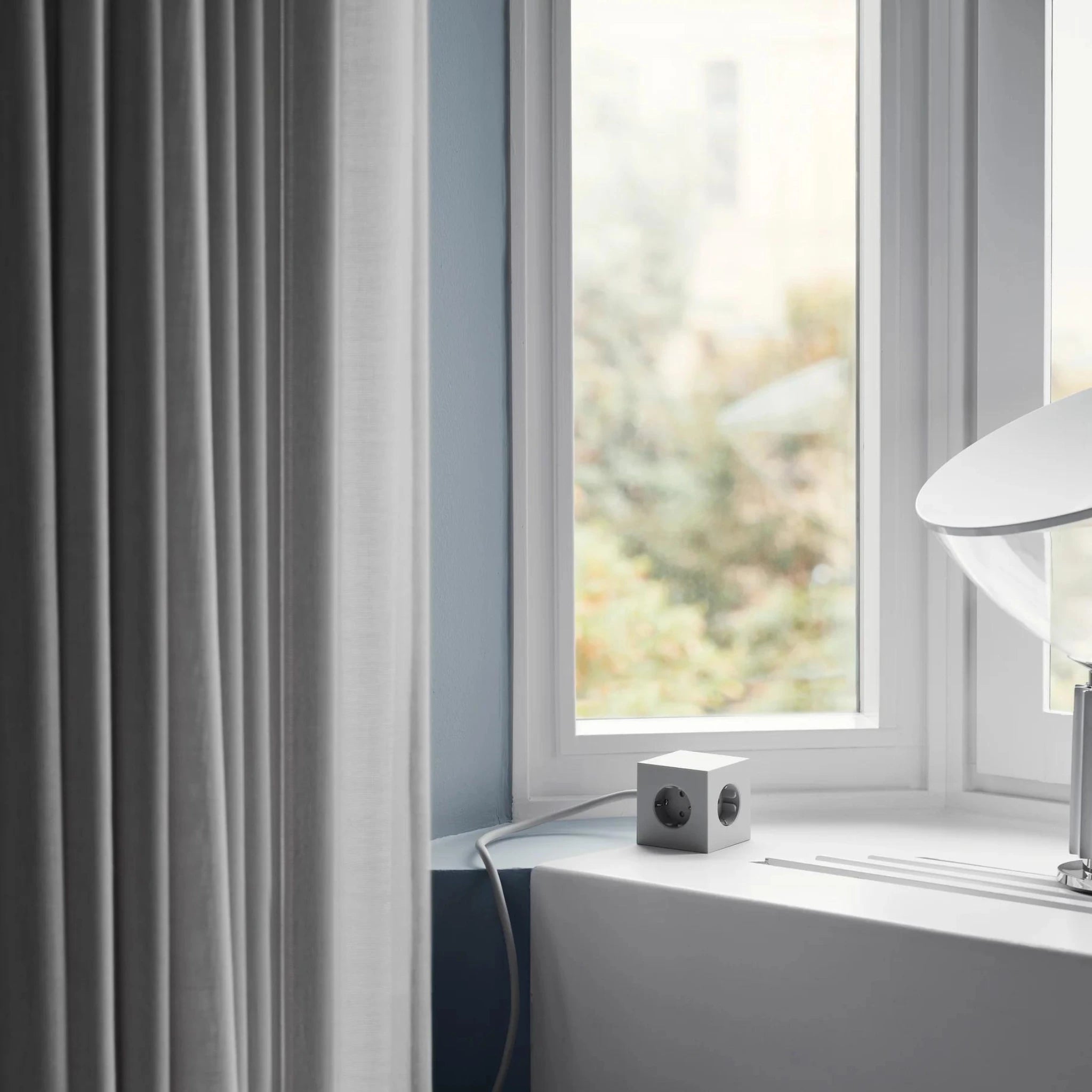 Een kleine witte beveiligingscamera staat op een vensterbank naast een moderne witte lamp en een Avolt Square 1 USB-C Stekkerdoos Gotland Grijs (1,8 m), met dunne gordijnen en vaag groen zichtbaar door het raam.