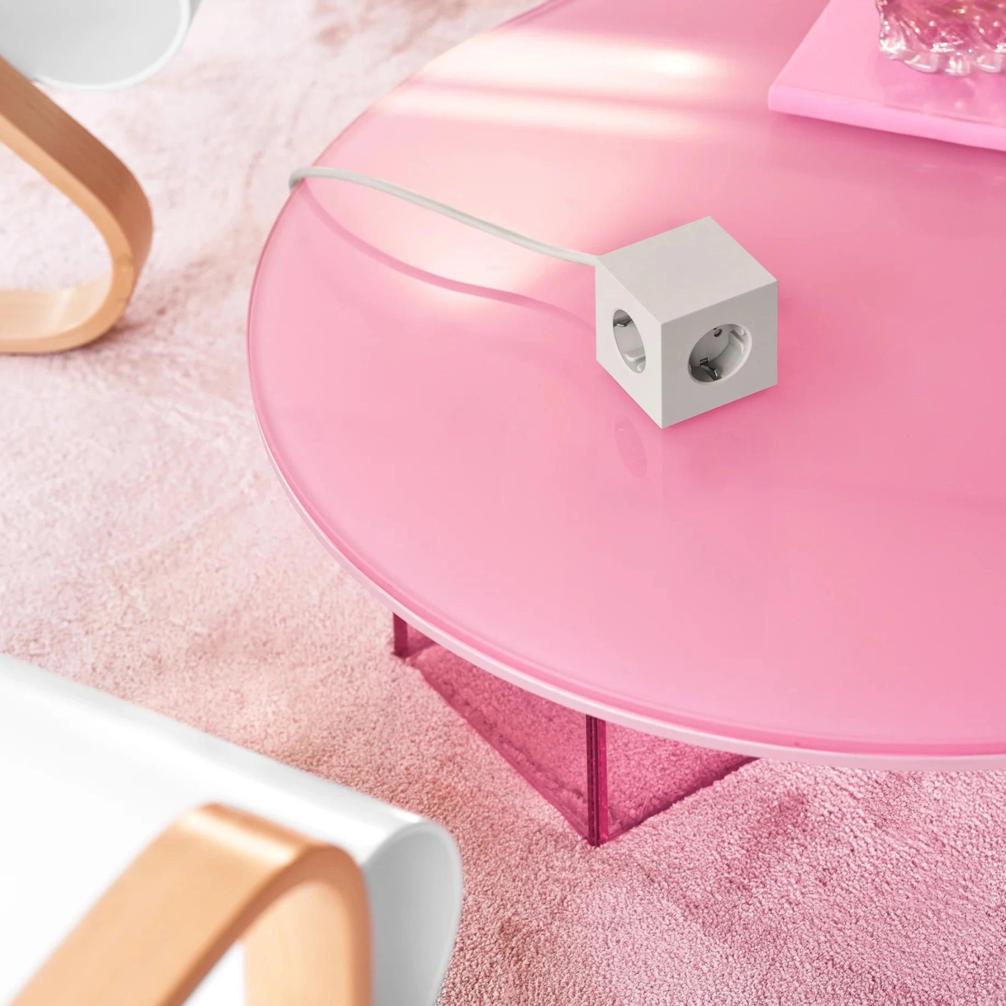 Een Avolt Square 1 USB-C Stekkerdoos Gotland Grijs 1,8 m staat op een glanzend roze ronde tafel in de buurt van een houten fauteuil en gestructureerd tapijt en voegt een modern tintje toe aan de ruimte.