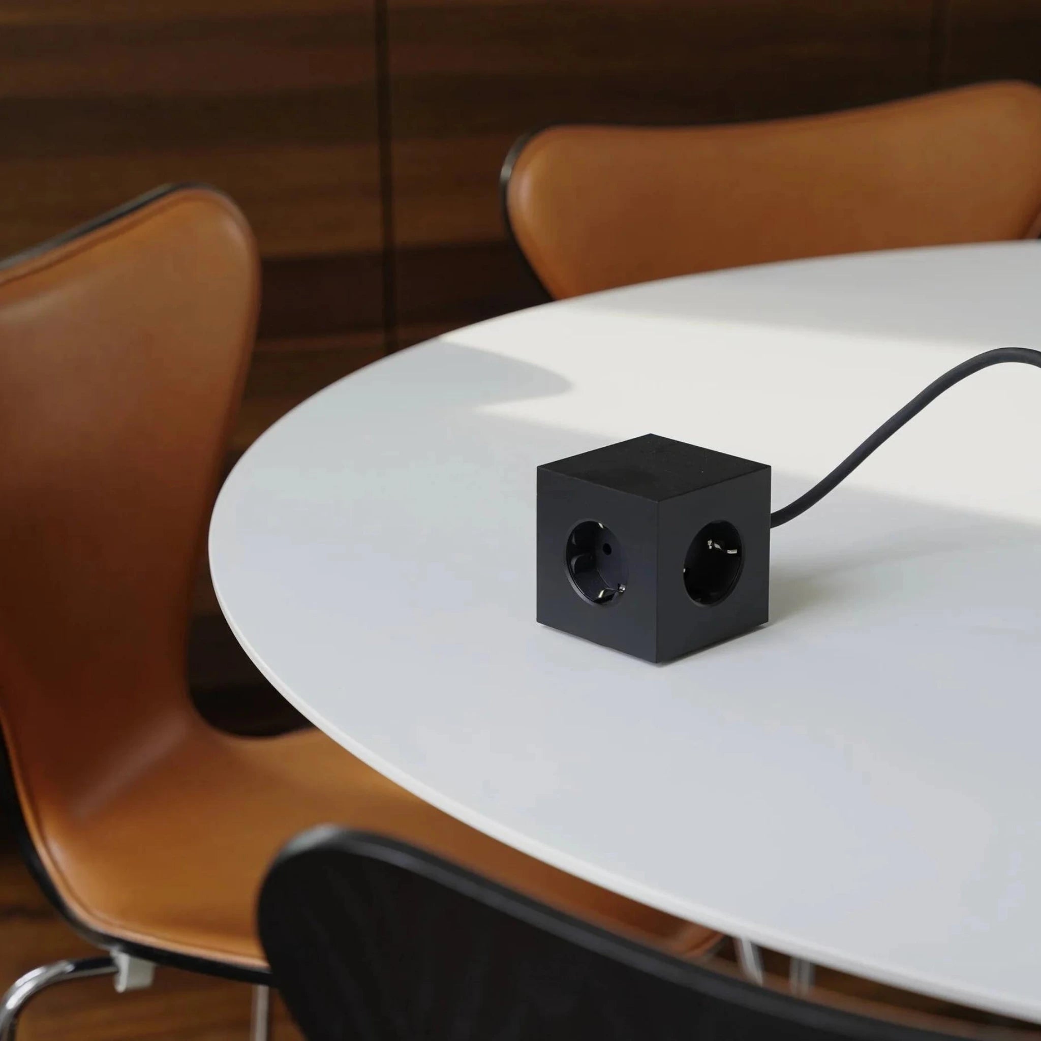 Een Avolt Square 1 USB-C Stekkerdoos Stockholm Zwart van 1,8 m van Avolt rust op een ronde witte tafel, omringd door bruine en zwarte moderne stoelen in een kamer met houten muren.