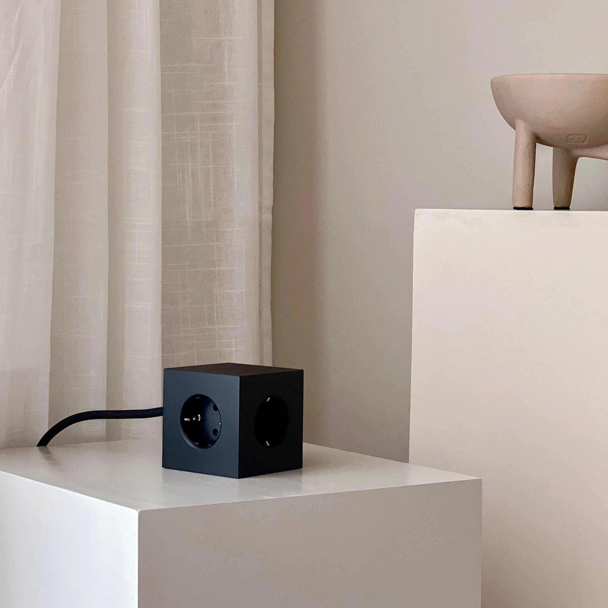 Een Avolt Square 1 USB-C Stekkerdoos Stockholm Zwart 1,8 m staat op een witte sokkel bij een beige muur en een gordijn. Een beige driepootkom is gedeeltelijk zichtbaar op een andere sokkel in het minimalistische, moderne tafereel.