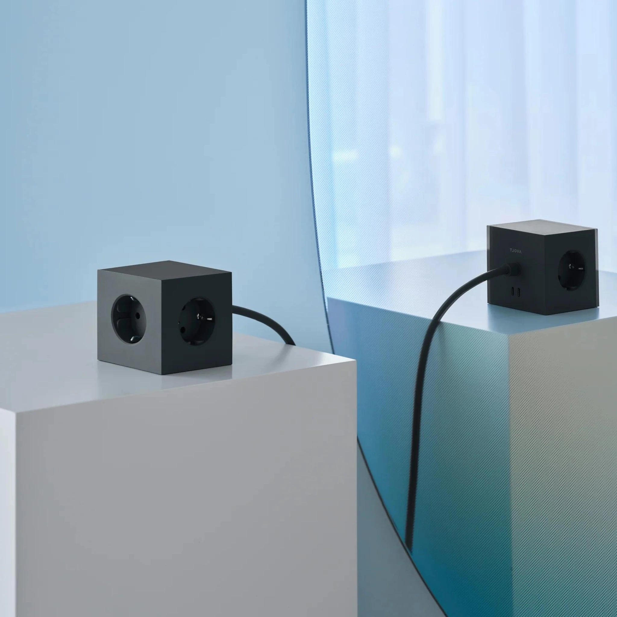 Een Avolt Square 1 USB-C Stekkerdoos Stockholm Zwart 1,8 m stekkerdoos van Avolt met 3 stopcontacten rust op een witte sokkel, met het snoer erachter, gespiegeld in de buurt voor een strakke, minimalistische look.