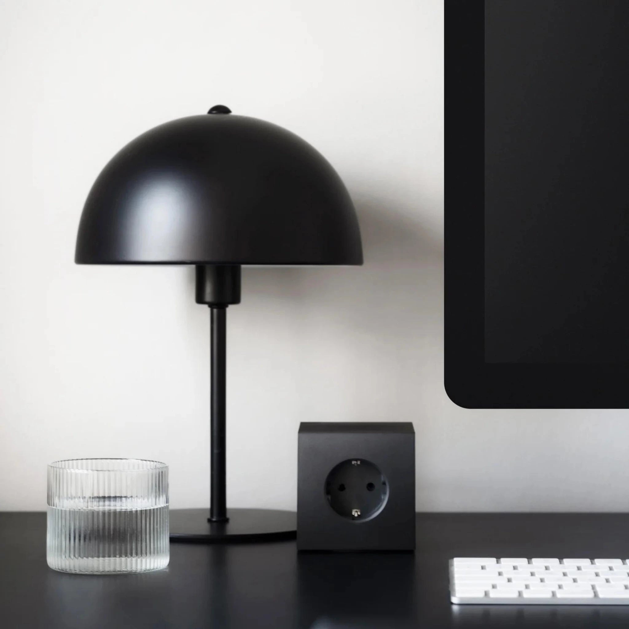 Een moderne zwarte bureaulamp, een glas water, een Avolt Square 1 USB-C Stekkerdoos Stockholm Zwart 1,8 m van Avolt en een deel van een computermonitor en toetsenbord staan opgesteld op een zwart bureau tegen een lichtgekleurde muur.